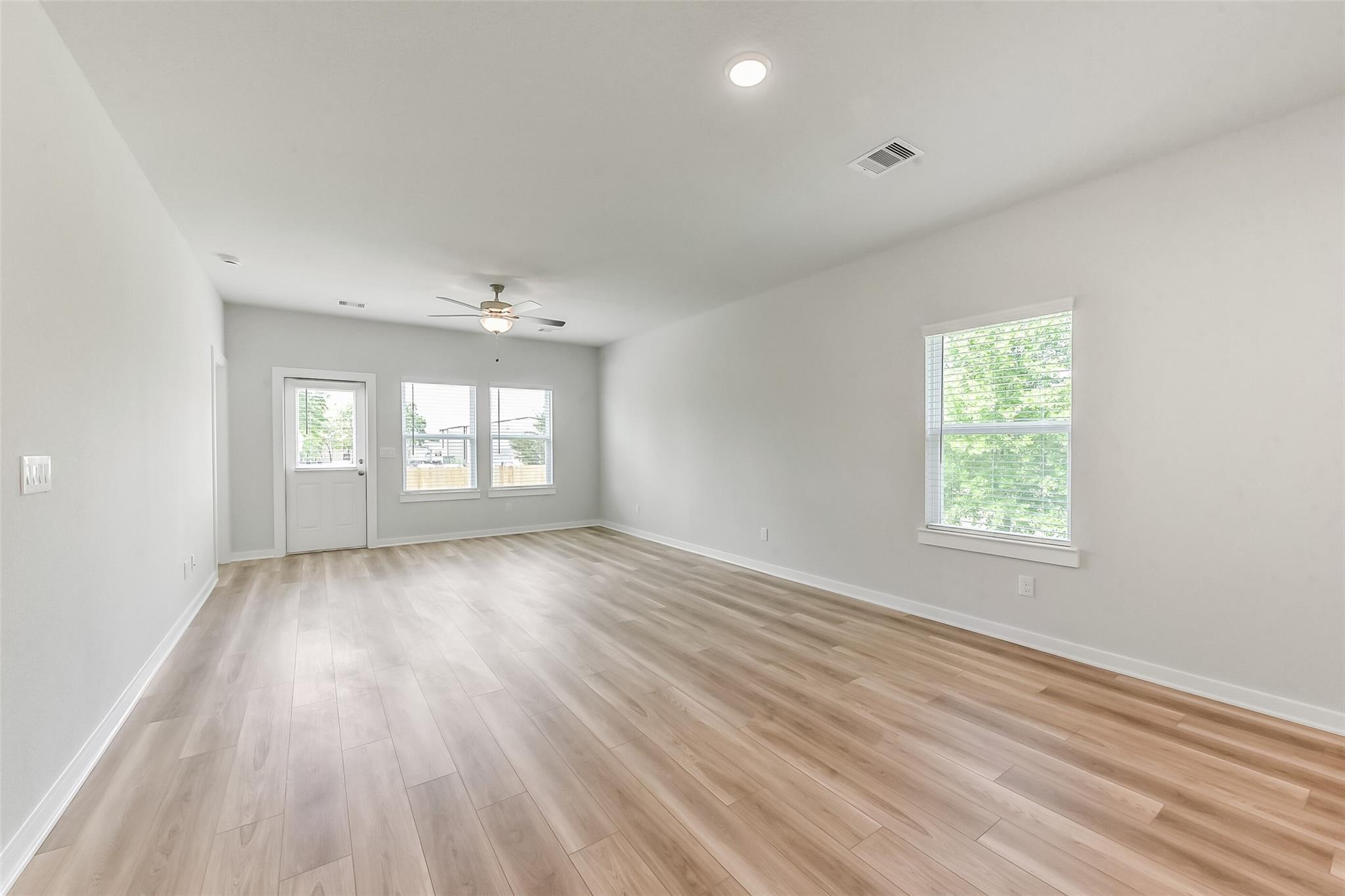 Spacious empty living room with light wood laminate floors, gray walls, ceiling fan, and large windows in Davidson Homes The Frio G, Magnolia TX