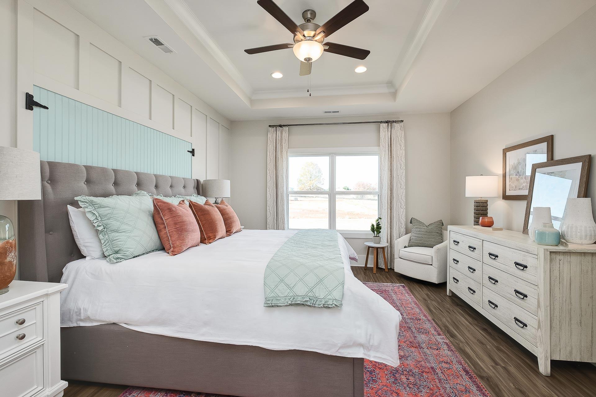 Spacious master bedroom at Newby Chapel in Athens AL with tufted gray bed, teal pillows, hardwood floors, accent chair, and ceiling fan