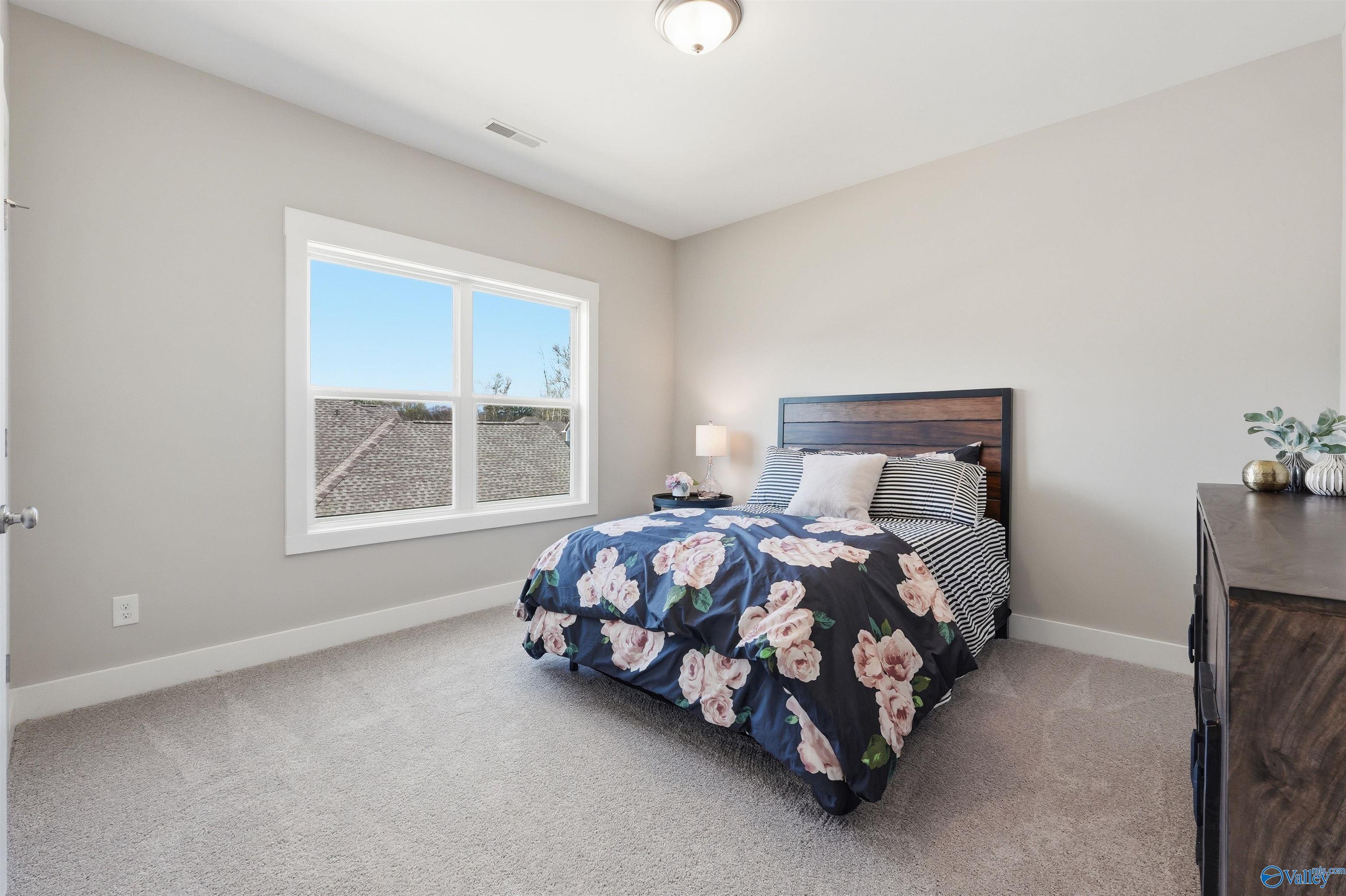 Cozy bedroom with navy floral bedding, wooden headboard, nightstand lamp, and large sunny window in Evermore Homes Oxford B, Owens Cross Roads AL