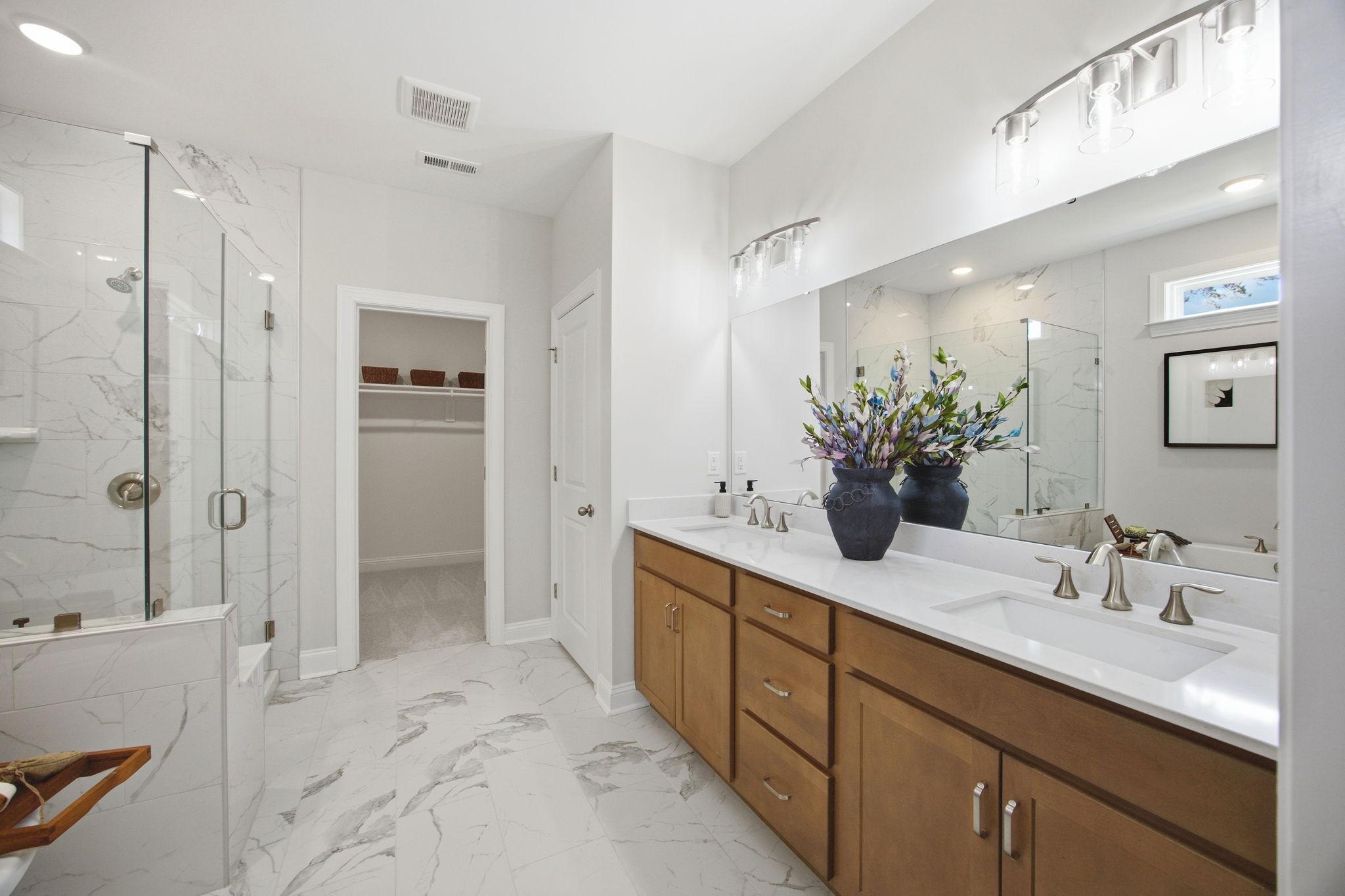 Modern primary bathroom at Enclave at Belmont with dual wood vanities, marble-look floors, and a glass-enclosed shower.