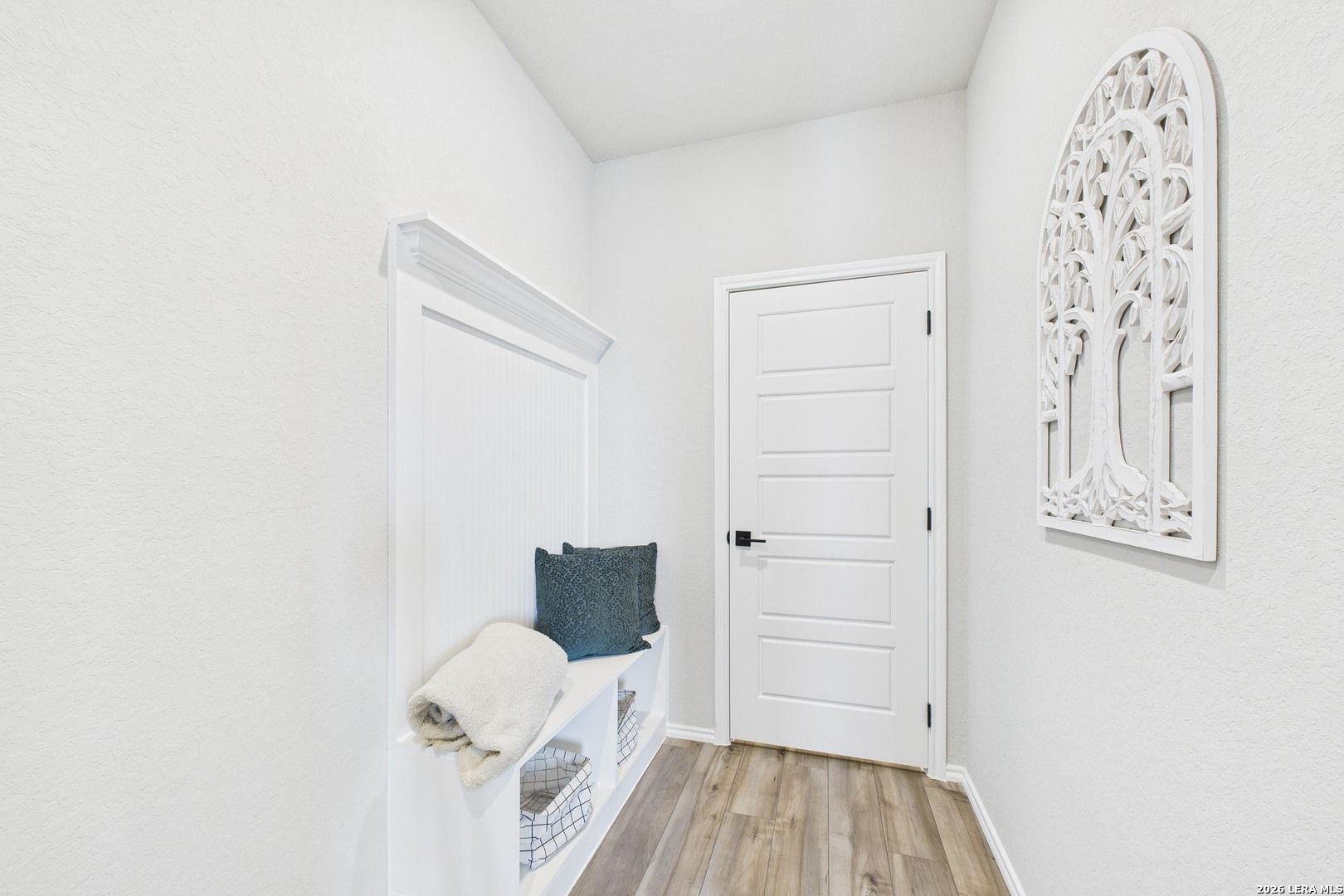 Bright white entryway with built-in bench, shelves, gray cushion, tree wall art, and wood floors in Davidson Homes The Douglas E, Bricewood, San Antonio