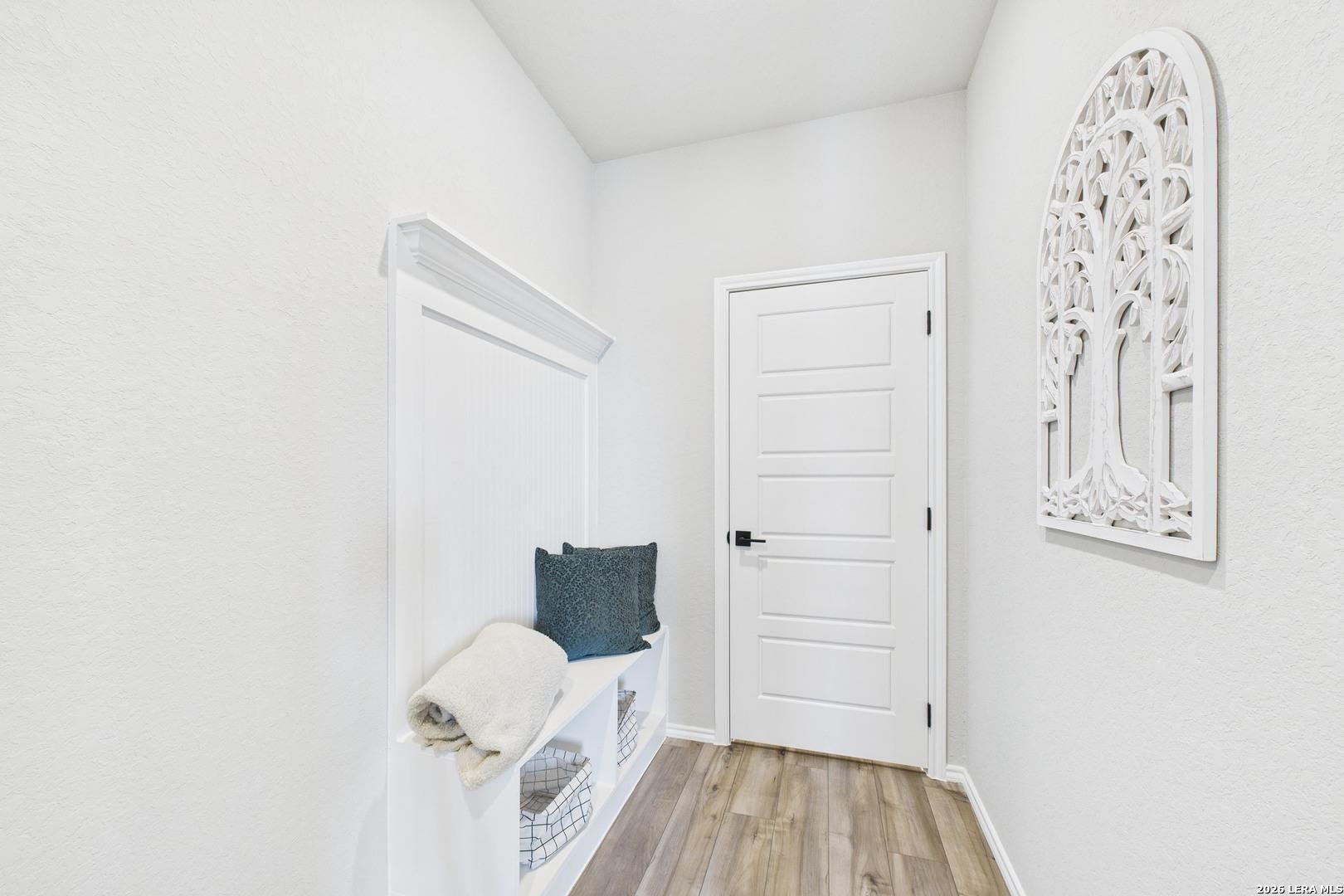 Bright white entryway with built-in bench, shelves, gray cushion, tree wall art, and wood floors in Davidson Homes The Douglas E, Bricewood, San Antonio