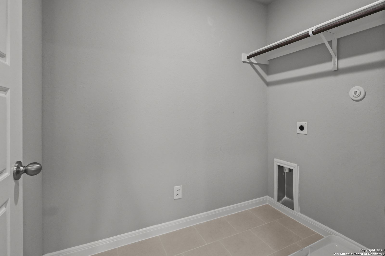 Functional laundry room with gray walls, wooden utility shelf, washer-dryer hookups, and beige tile floor in Davidson Homes The Gillian C, San Antonio