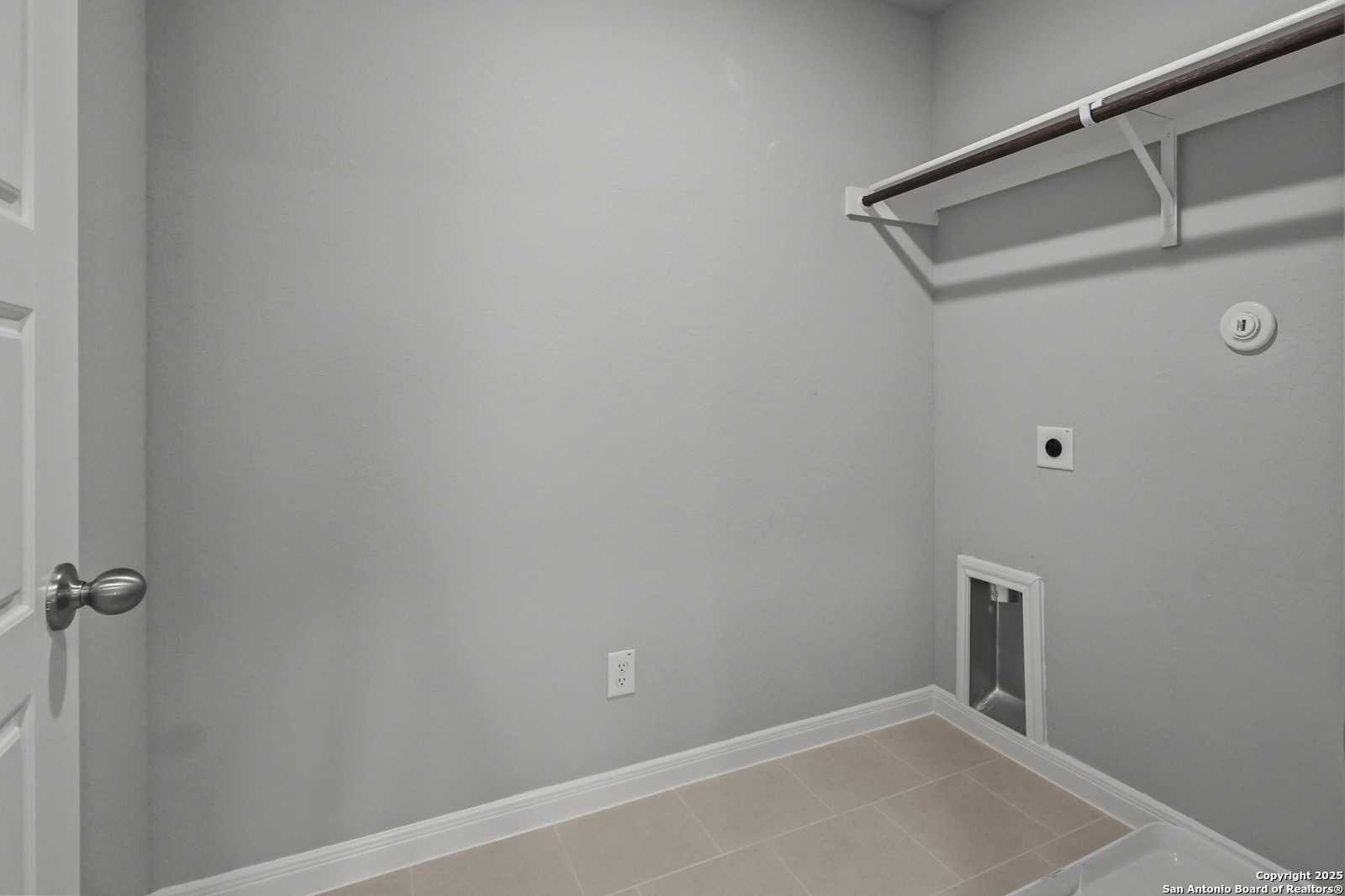 Functional laundry room with gray walls, wooden utility shelf, washer-dryer hookups, and beige tile floor in Davidson Homes The Gillian C, San Antonio