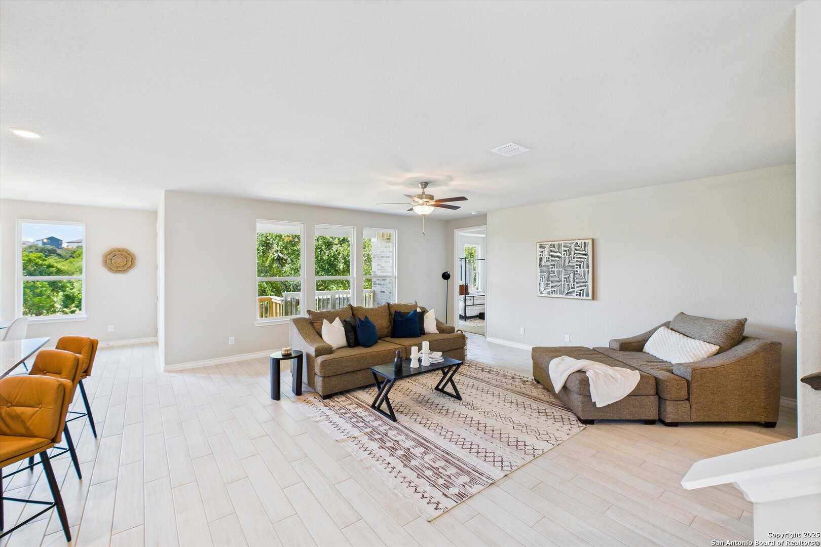 Bright open living room with large windows, neutral walls, brown sofa, ceiling fan, and rug in The Jennings H home, Ladera, San Antonio