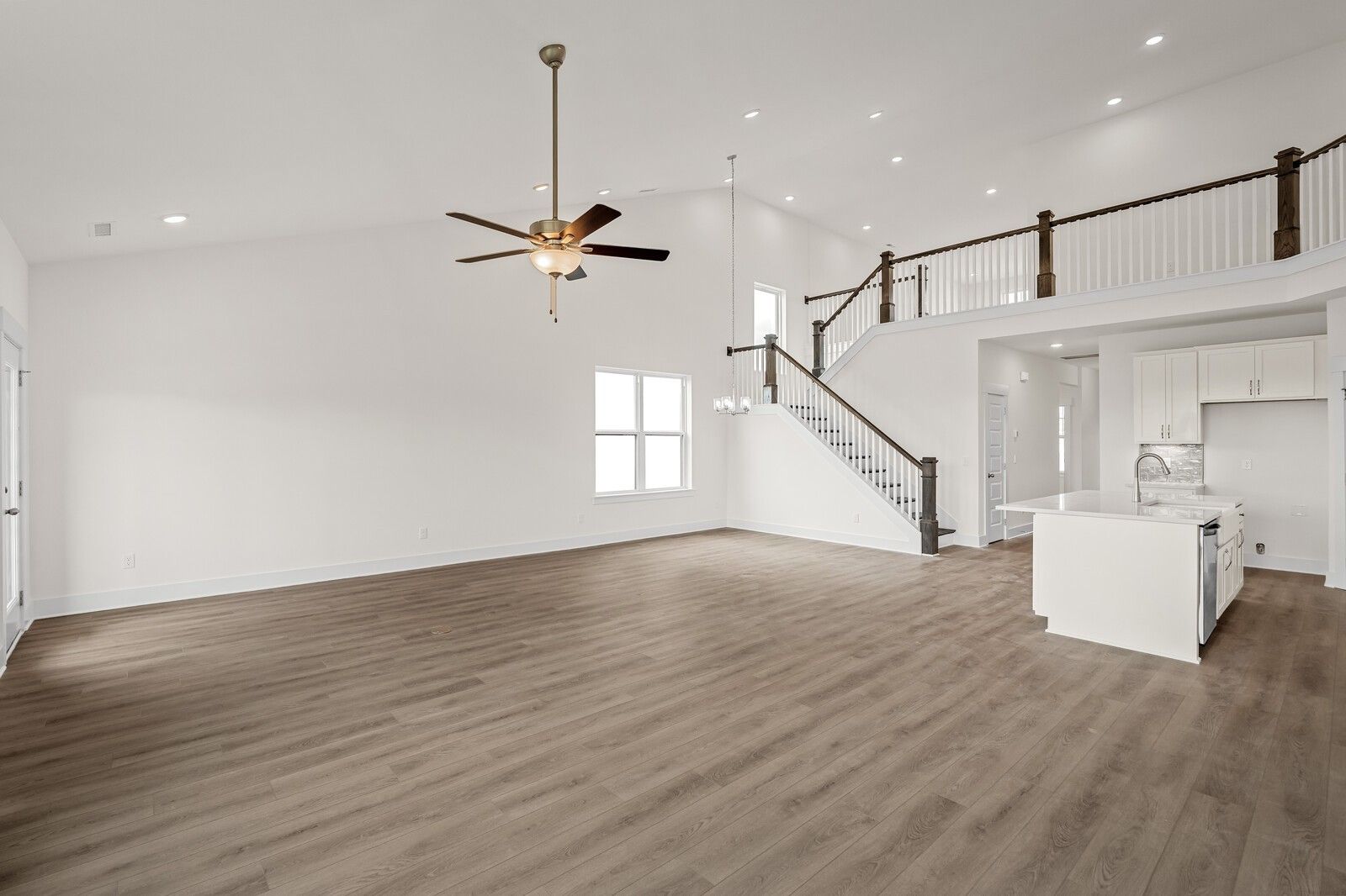 Spacious open living area with hardwood floors, white kitchen island, ceiling fan, and wooden staircase to balcony in The Ridgeport D, Mt. Juliet, TN