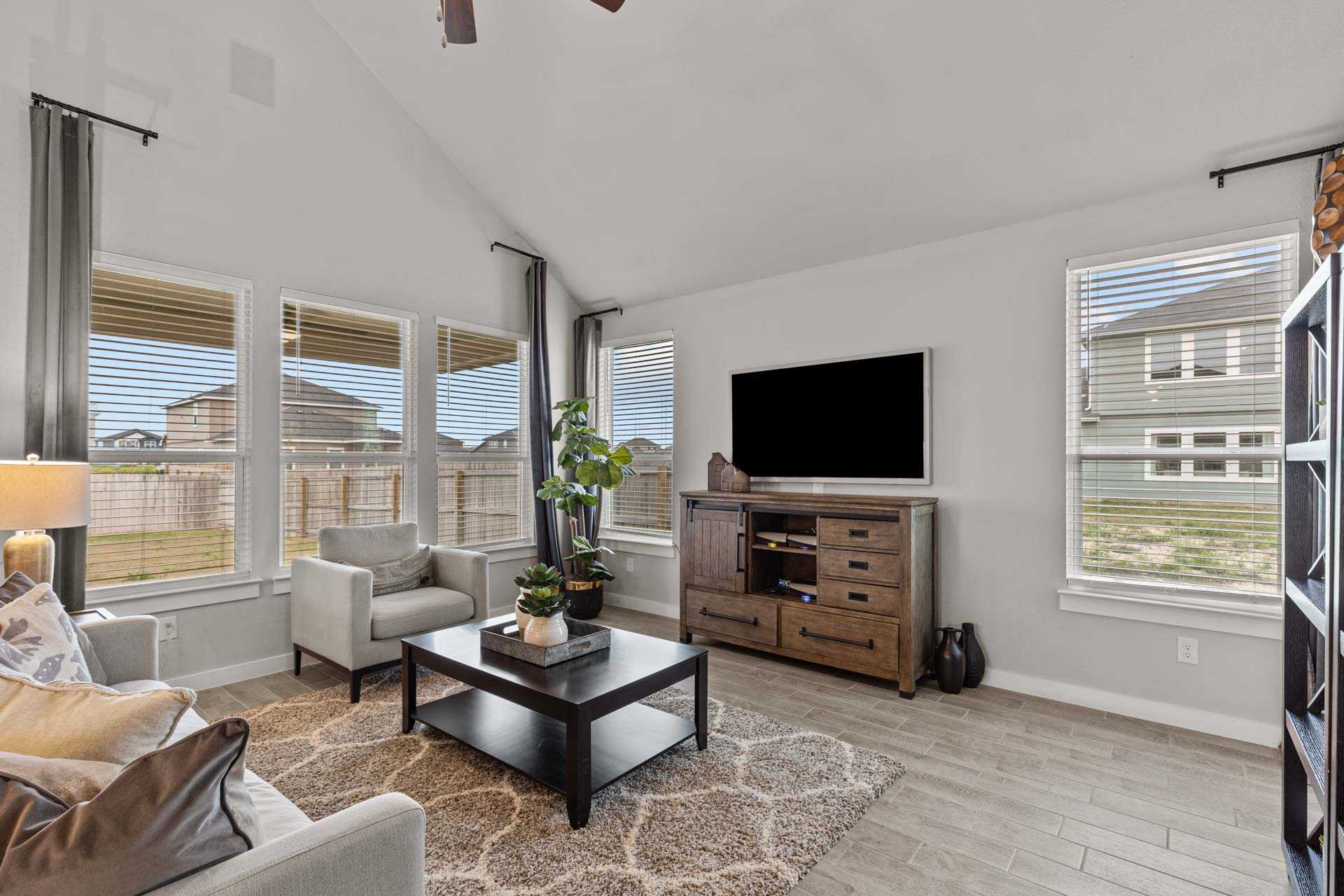 Spacious living room in The Collin B featuring vaulted ceiling, large windows, wooden entertainment center, and cozy gray seating