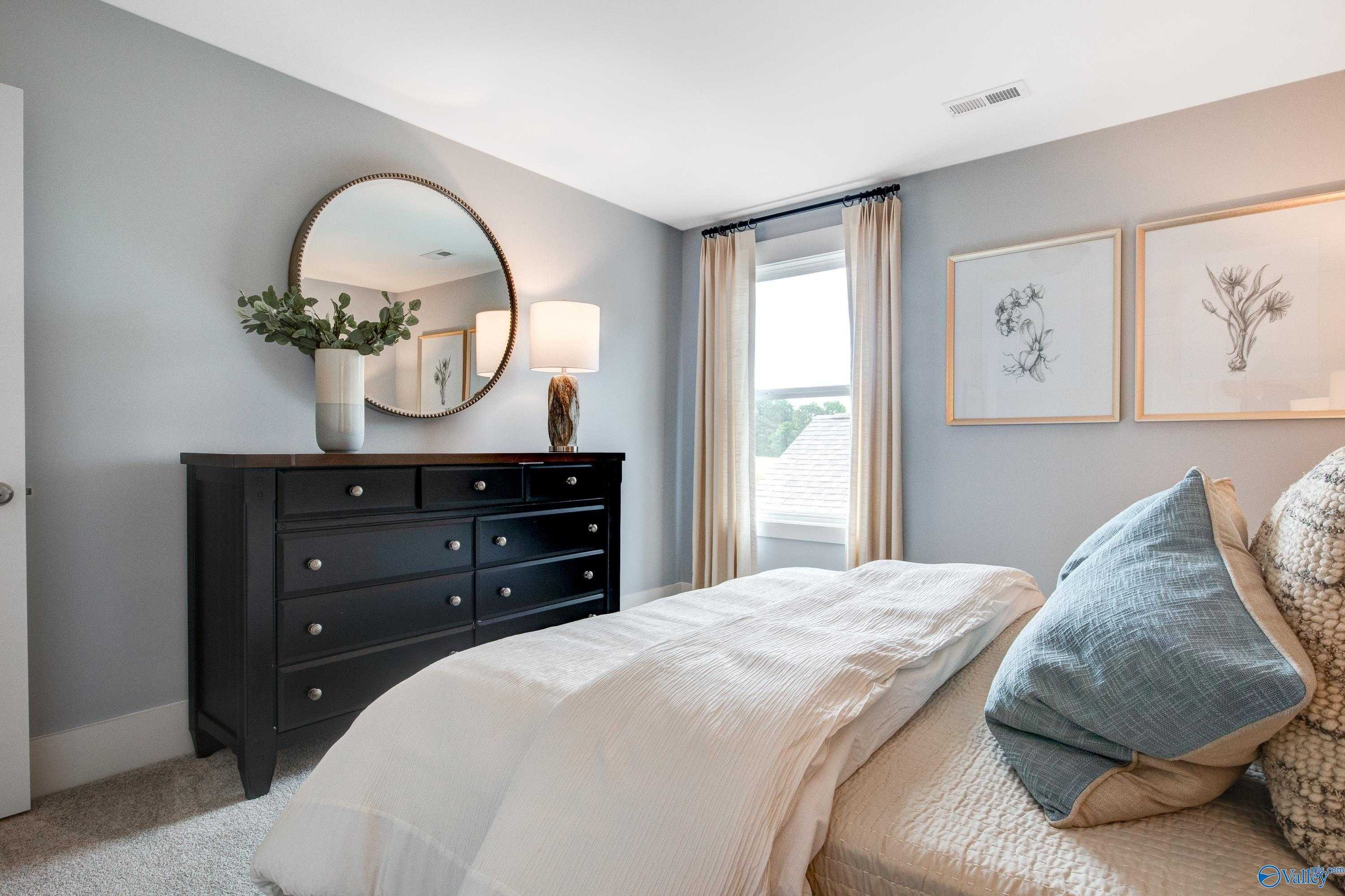 Cozy bedroom with light gray walls, round mirror over dark wood dresser, white bed with blue pillows, and botanical art in Davidson Homes The Covington C, Decatur, Alabama