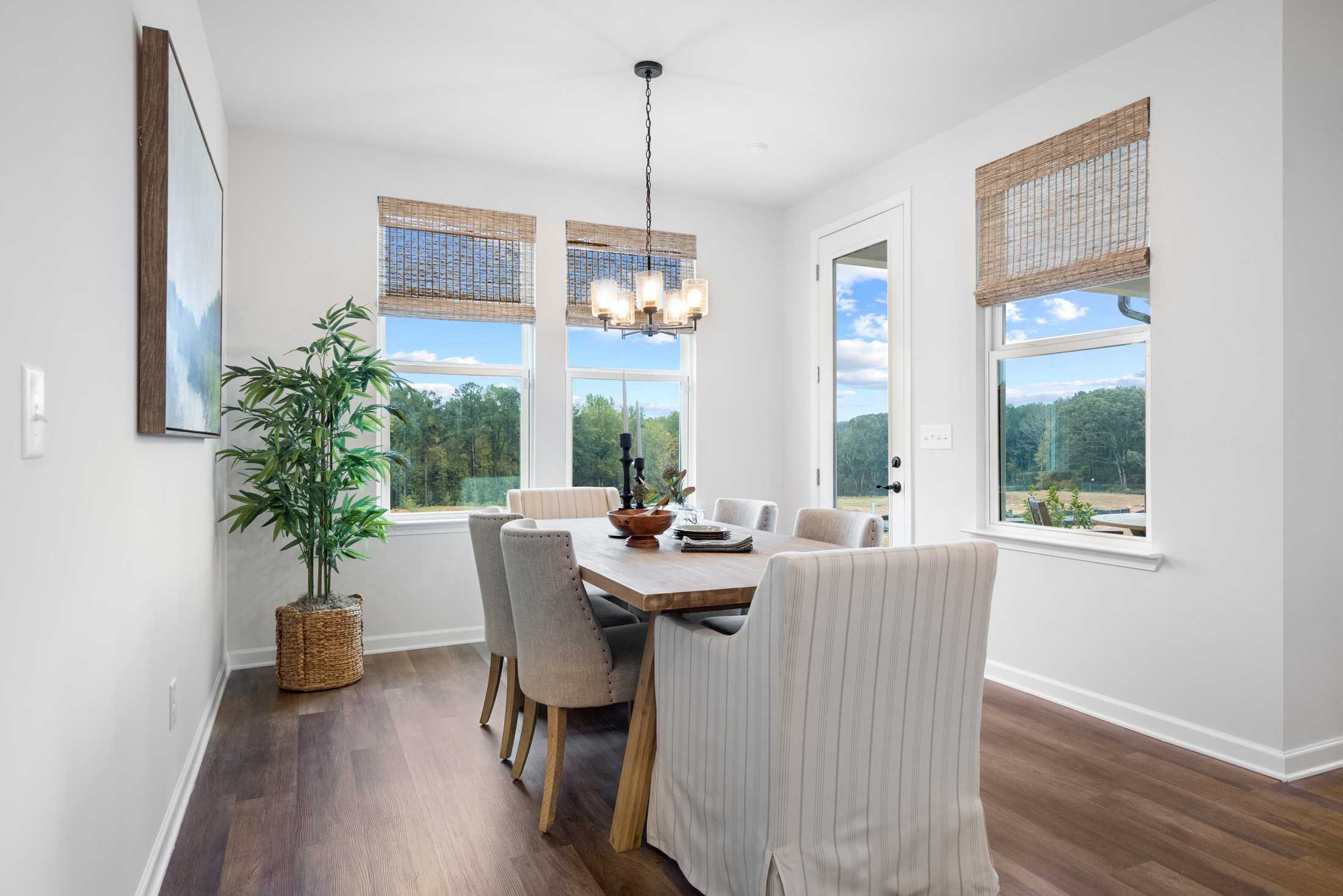 Spacious dining room in The Edison C with wooden table, slipcovered chairs, chandelier, potted plant, and large windows overlooking greenery