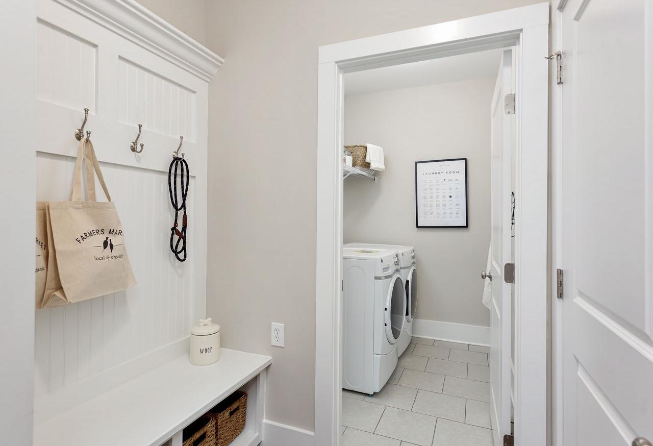 Spacious mudroom in The Rockford home design with white paneled bench, hooks, canvas bag, dog leash, and adjacent laundry room featuring washer dryer