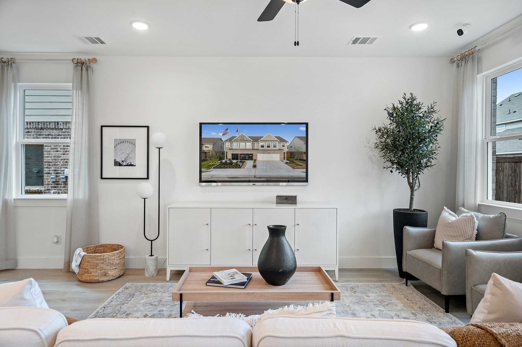 Cozy modern living room with wall-mounted TV showing home exterior, beige sofa, potted olive tree, and sheer curtains in Davidson Homes The Wake D, Wylie, Texas
