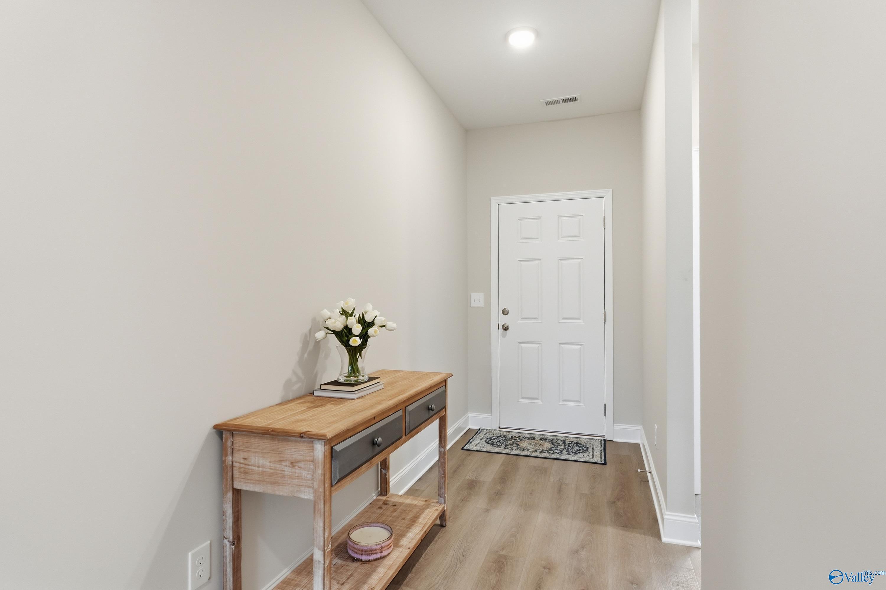 Elegant hallway with wooden console table, white roses vase, and rug leading to white door in The Asheville 3-bedroom home, Toney, Alabama