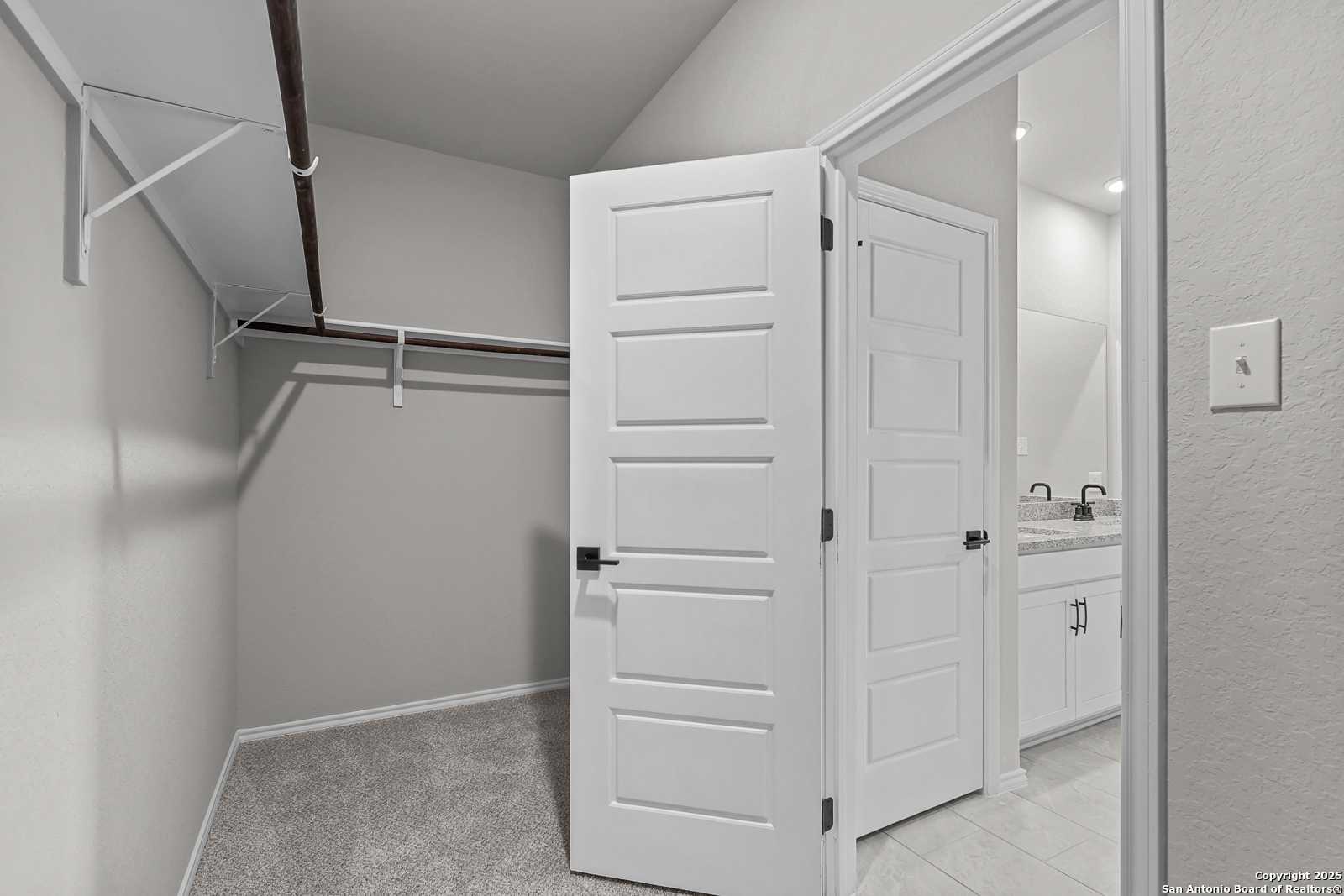 Spacious walk-in closet with gray walls, carpet floor, open white doors, built-in rods and shelves adjacent to master bath vanity in Davidson Homes The Daphne K, San Antonio