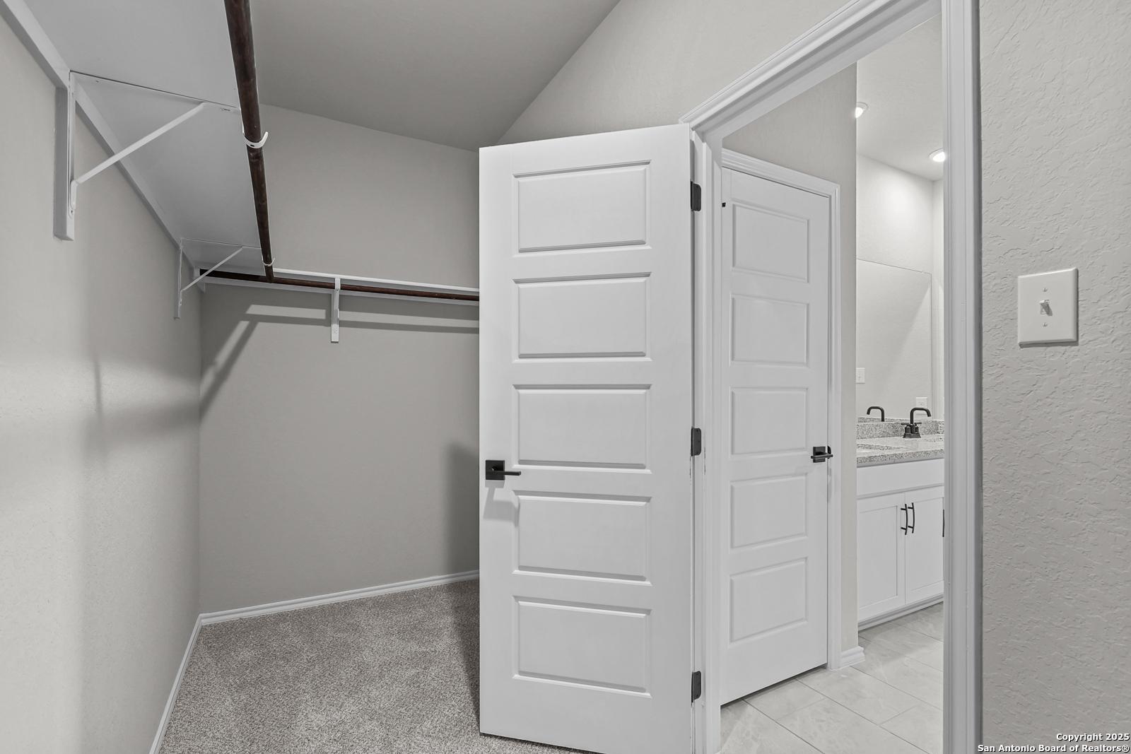 Spacious walk-in closet with open double doors, hanging rods, and gray carpet adjacent to modern bathroom with double vanity in Davidson Homes The Daphne K, San Antonio