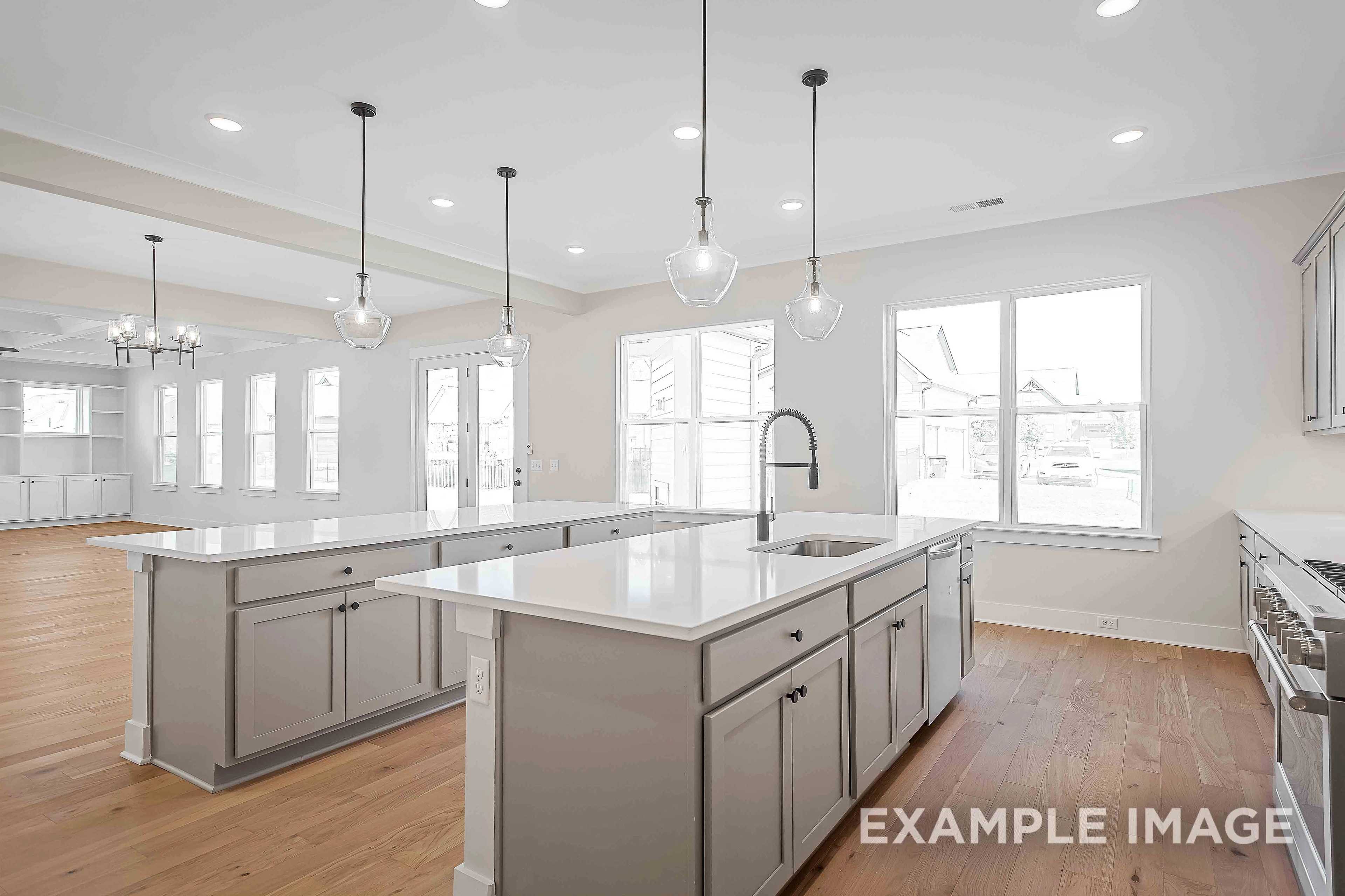 Spacious Alston A kitchen by Davidson Homes featuring large white island with sink, gray shaker cabinets, pendant lights, and open layout with hardwood floors