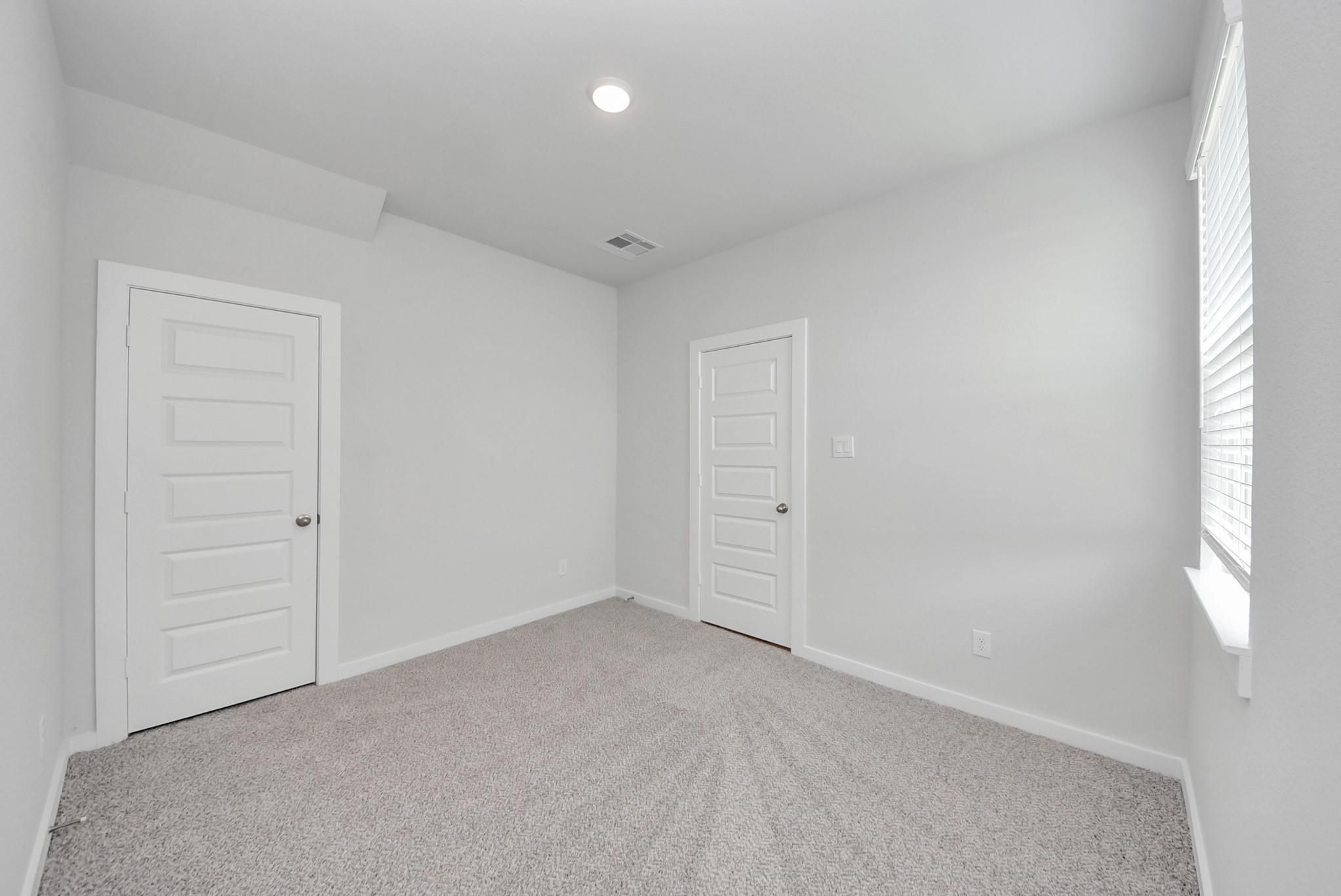 Bright empty bedroom with white doors, carpet flooring, and window with blinds in Davidson Homes San Marcos E plan, Beasley, TX