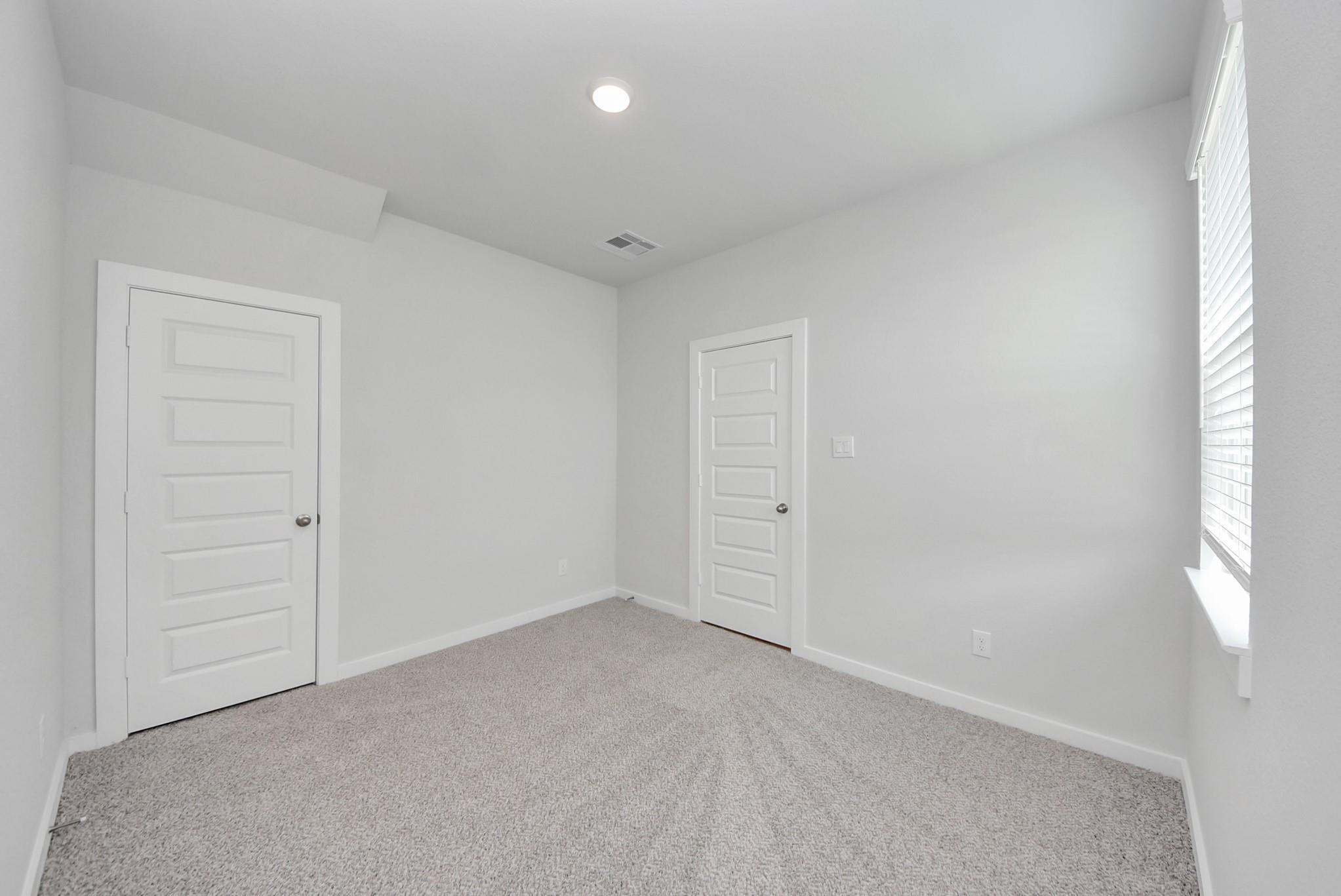 Bright empty bedroom with white doors, carpet flooring, and window with blinds in Davidson Homes San Marcos E plan, Beasley, TX
