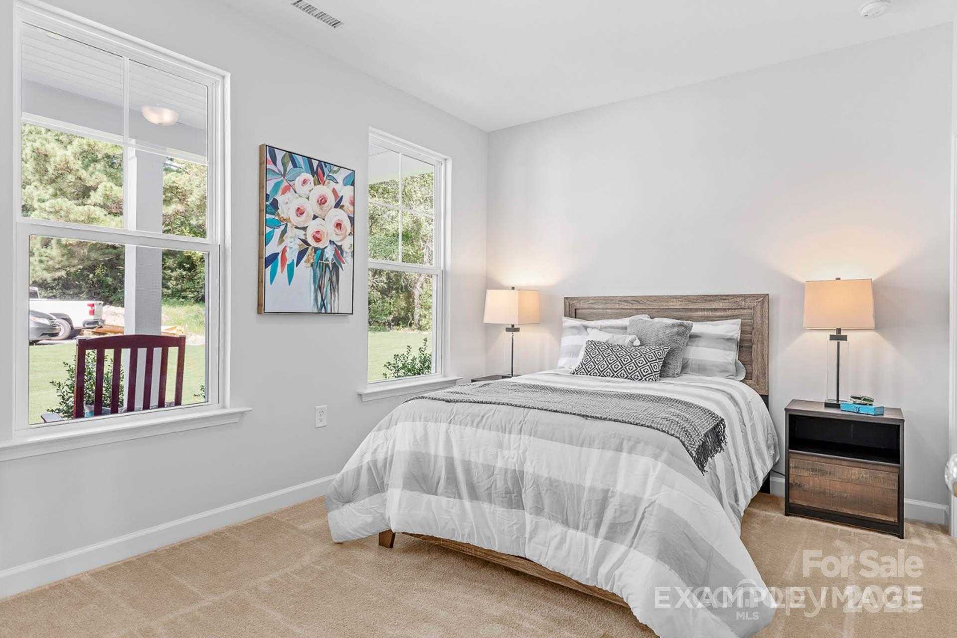 Bright bedroom with large windows overlooking lush backyard, gray quilted bed, wooden headboard, lamps in Davidson Homes The Chestnut F, Belmont NC