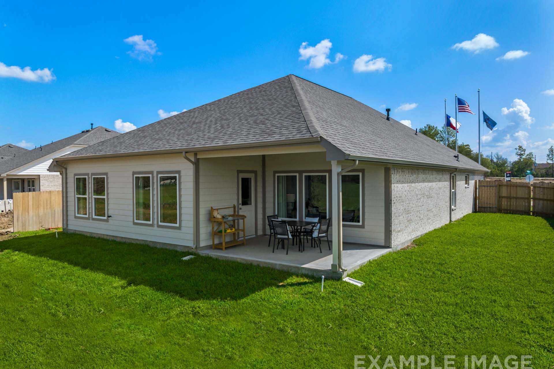 Rear view of The Acadia B single-story home featuring covered patio with wooden seating, US and Texas flags, and lush green backyard in Magnolia, Texas