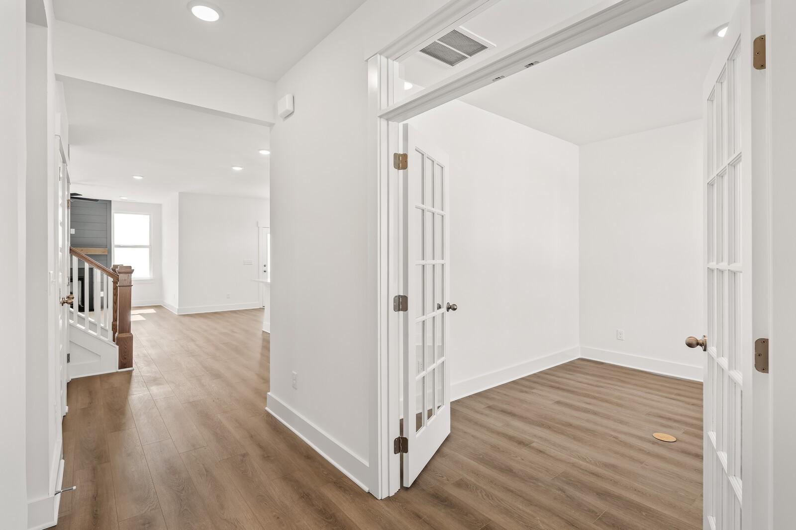 Bright hallway with open French doors to white study, hardwood floors, and wooden staircase in Davidson Homes The Ash A, Gallatin, TN