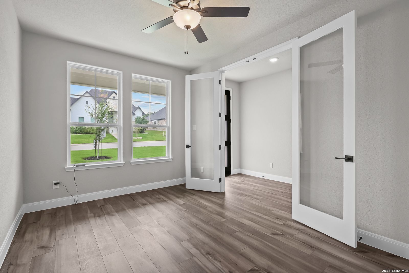 Bright bedroom with light gray walls, hardwood floors, ceiling fan, large windows, and open glass French doors in Davidson Homes The Belmont H, Castroville TX