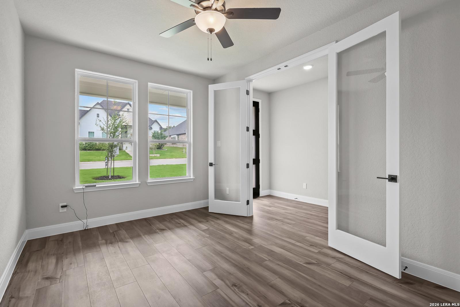Bright bedroom with light gray walls, hardwood floors, ceiling fan, large windows, and open glass French doors in Davidson Homes The Belmont H, Castroville TX