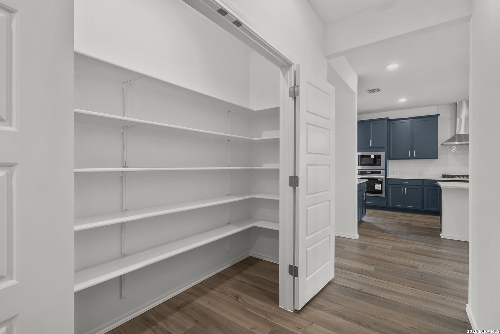 Spacious walk-in pantry with white shelving open to modern navy blue kitchen in Davidson Homes The Glenwood E, Elmendorf, Texas