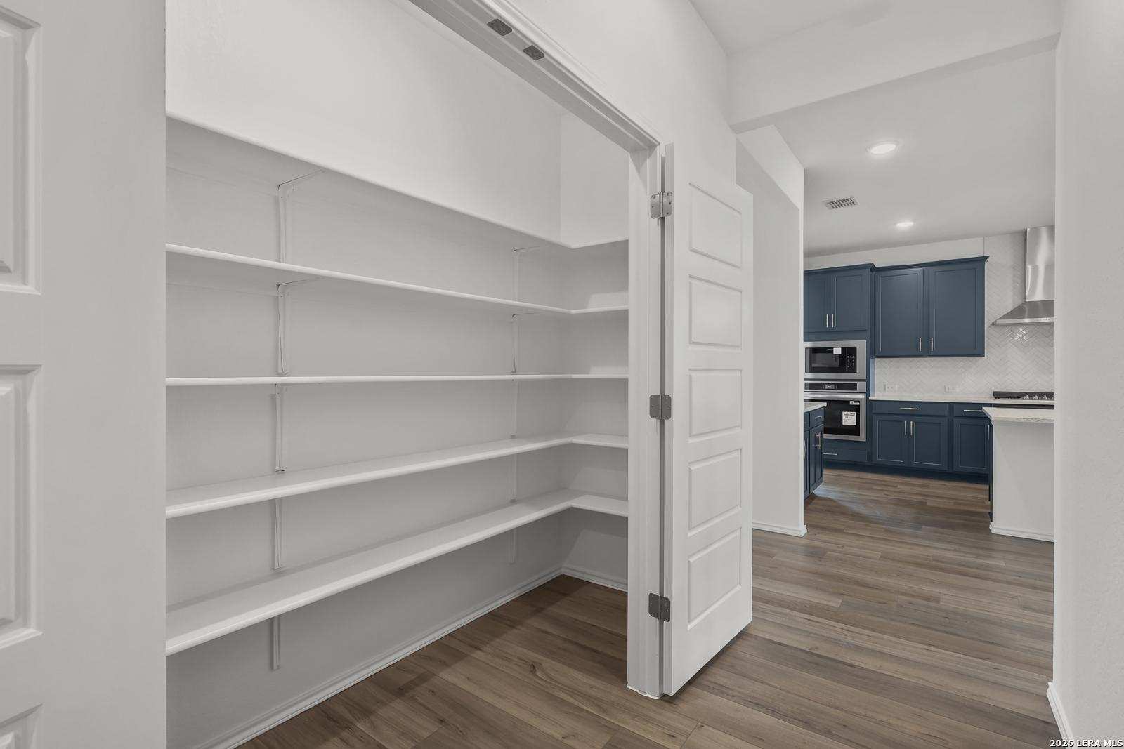 Spacious walk-in pantry with white shelving open to modern navy blue kitchen in Davidson Homes The Glenwood E, Elmendorf, Texas