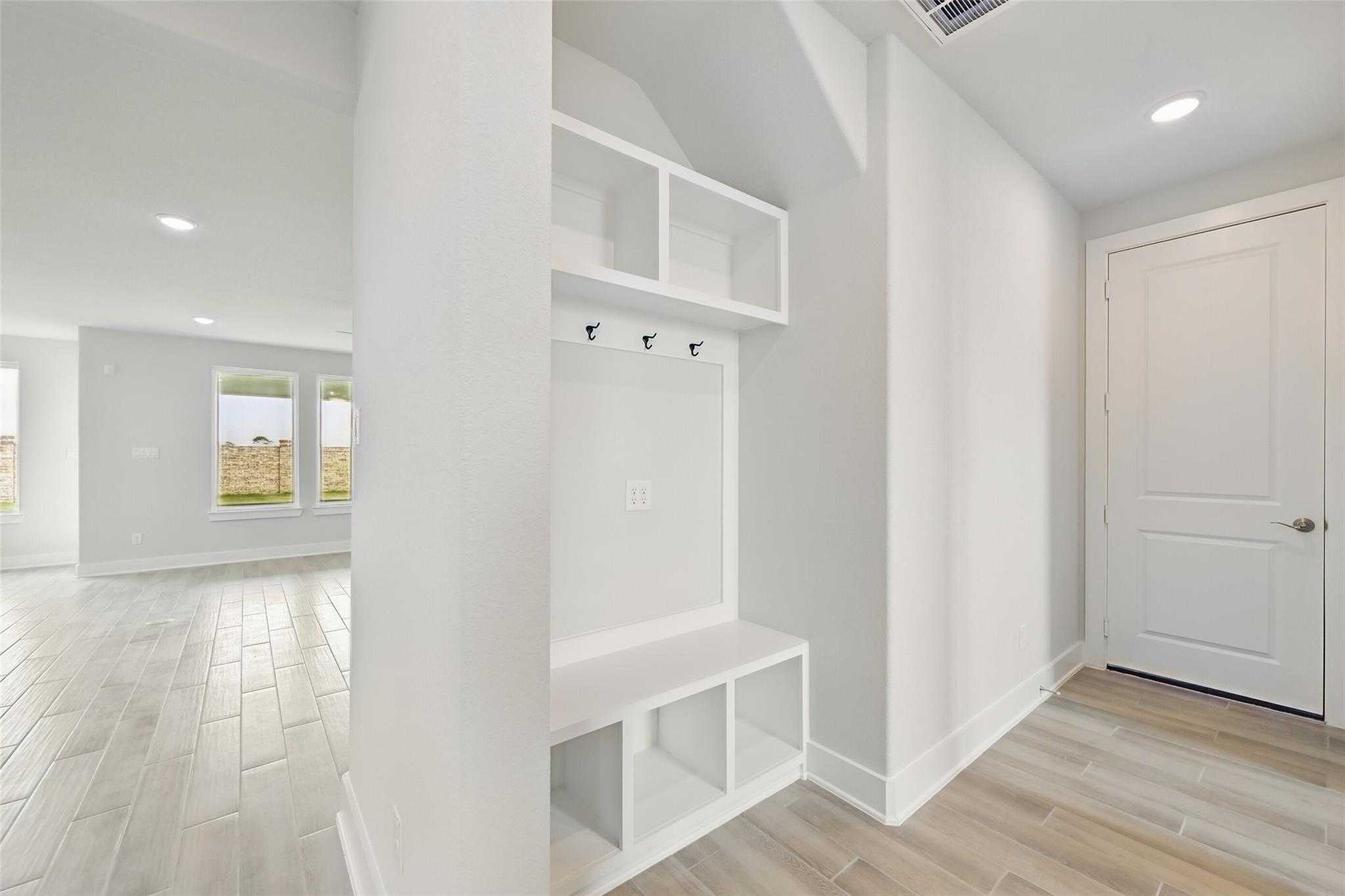 Spacious white mudroom with built-in shelves, bench, coat hooks, and light wood floors in Davidson Homes The Philip A, Lago Mar, Texas City