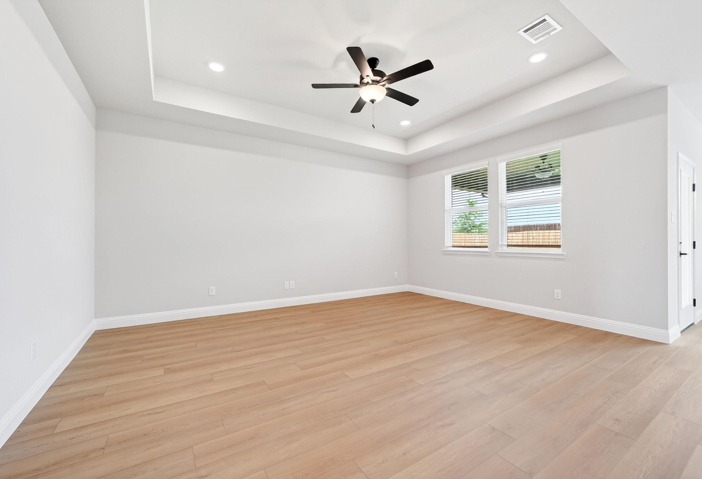 Spacious master bedroom in The Holly M featuring a tray ceiling, modern ceiling fan, and light wood-style flooring.