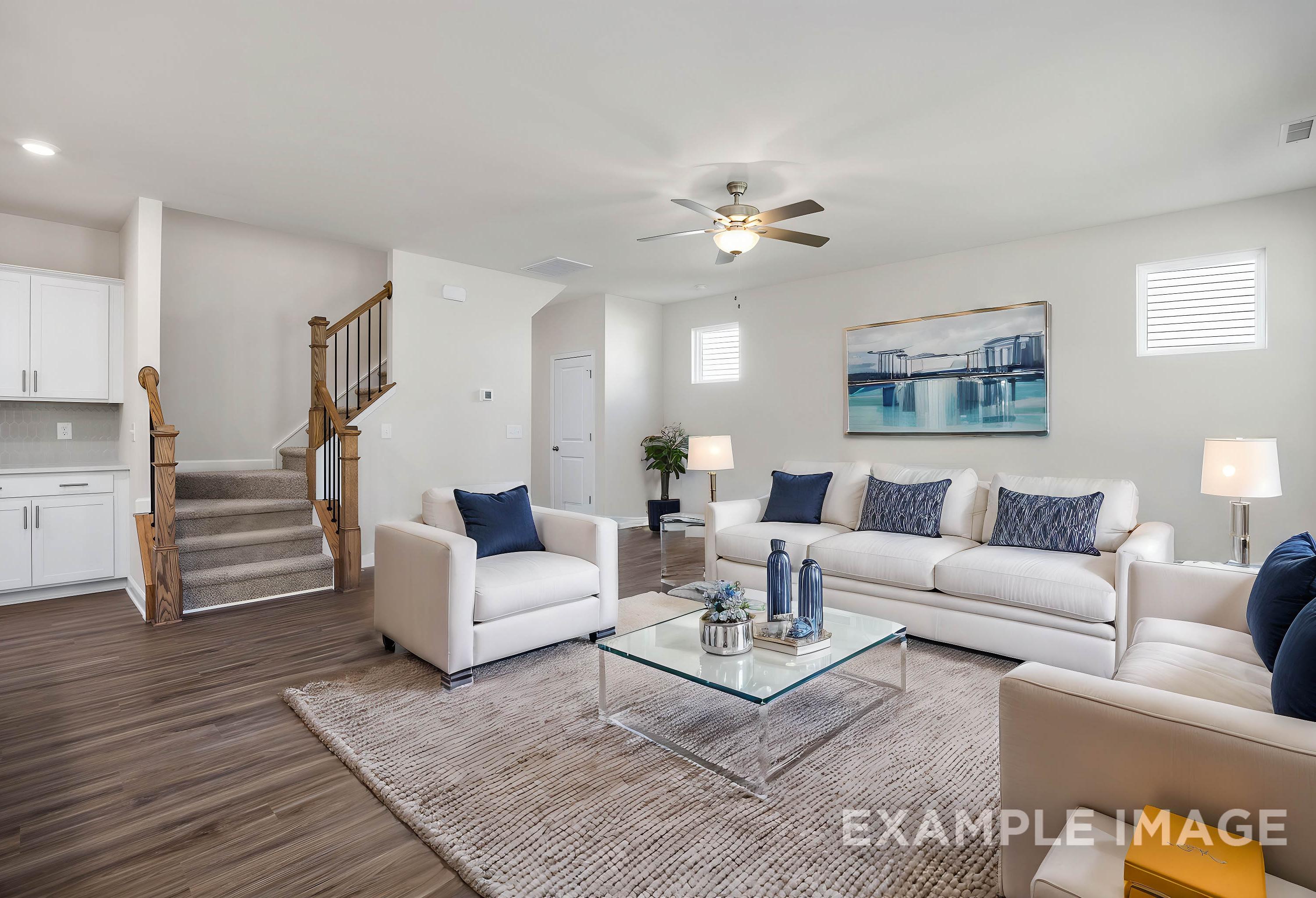Spacious living room in The Grace A 2-story home featuring white sectional sofas, blue pillows, glass coffee table, area rug, and open wooden staircase