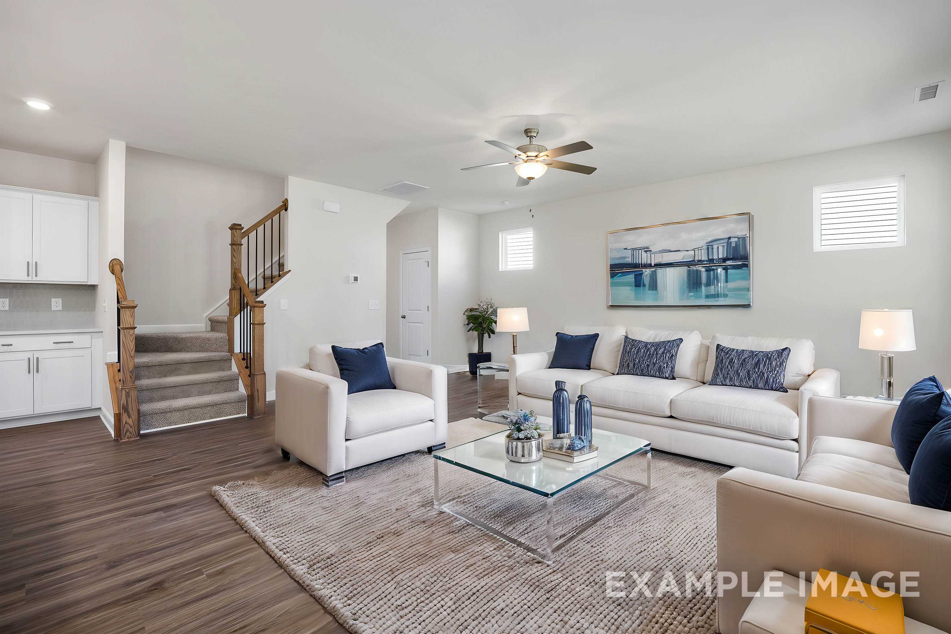Spacious living room in The Grace A 2-story home featuring white sectional sofas, blue pillows, glass coffee table, area rug, and open wooden staircase