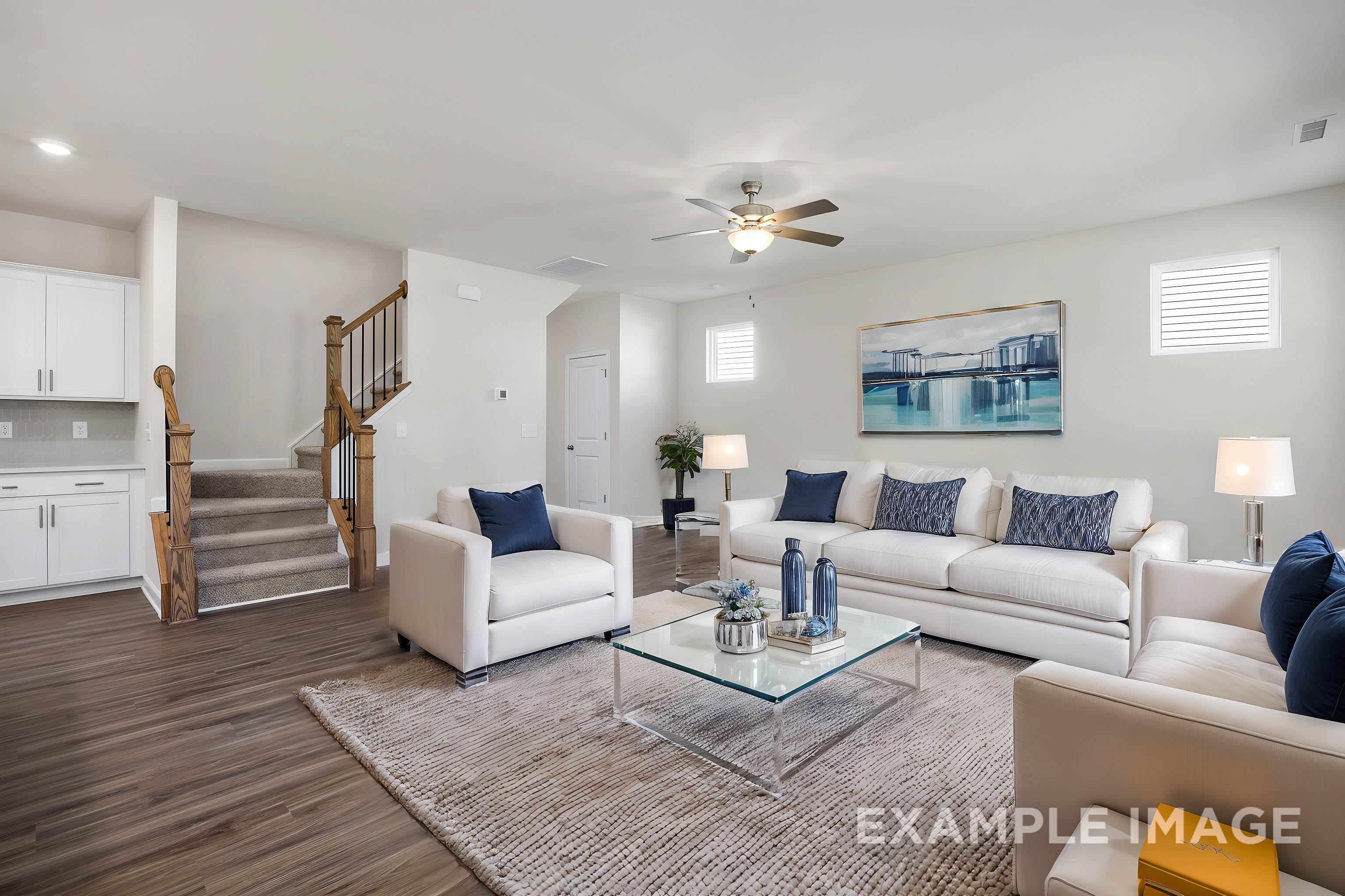 Spacious living room in The Grace A 2-story home featuring white sectional sofas, blue pillows, glass coffee table, area rug, and open wooden staircase