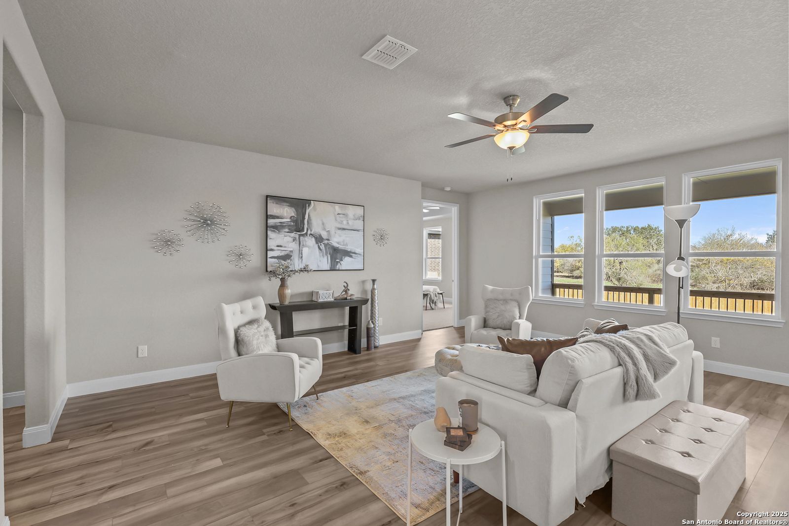 Cozy living room with beige sofa, white armchairs, hardwood floors, ceiling fan, and backyard views in The Jennings G, Castroville, Texas