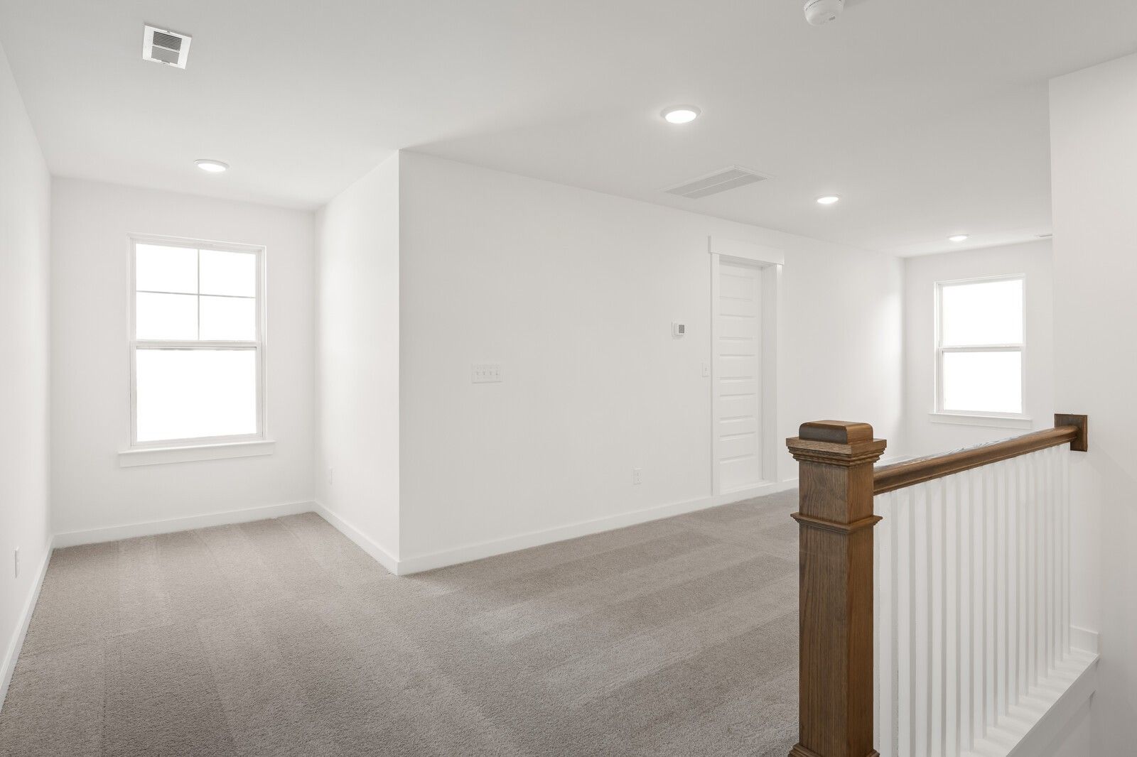 Bright upstairs hallway with white walls, gray carpet flooring, large windows, and wooden staircase railing in Davidson Homes The Ash A, Gallatin, TN