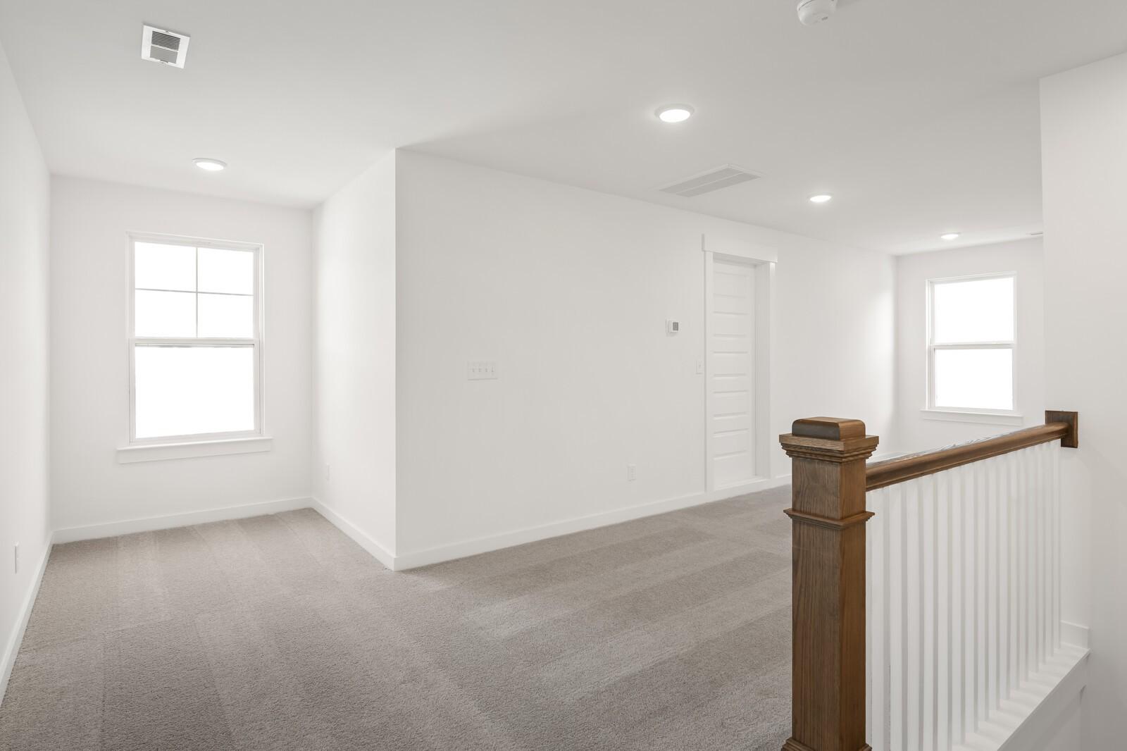 Bright upstairs hallway with white walls, gray carpet flooring, large windows, and wooden staircase railing in Davidson Homes The Ash A, Gallatin, TN