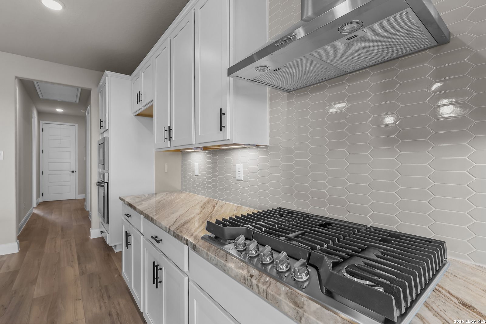 Gas cooktop with stainless hood, white cabinetry, and hexagon tile backsplash in Davidson Homes Foster E kitchen, Castroville, TX