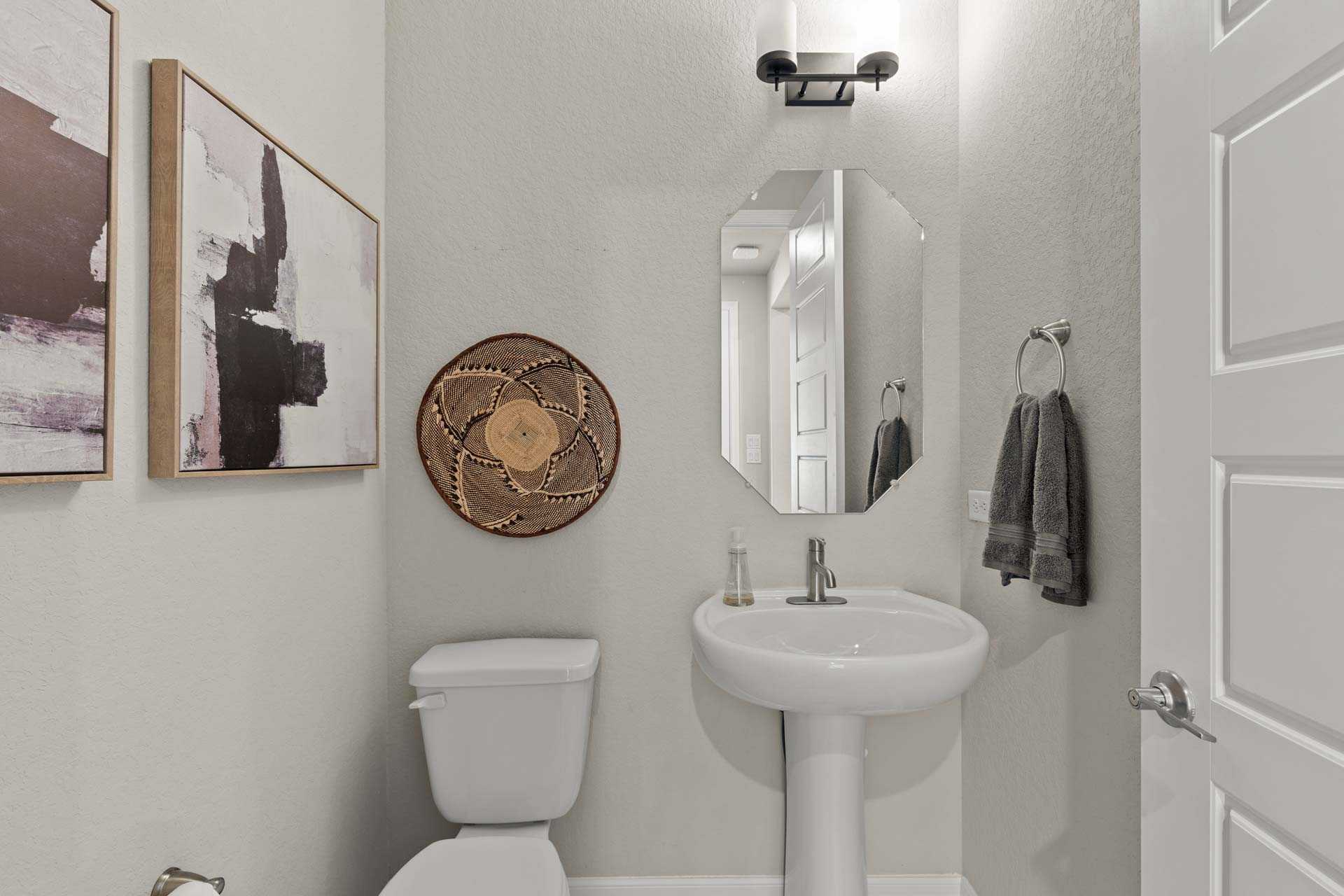 Spacious powder room in The Summerlin B with pedestal sink, oval mirror, abstract wall art, woven decor, and neutral tones