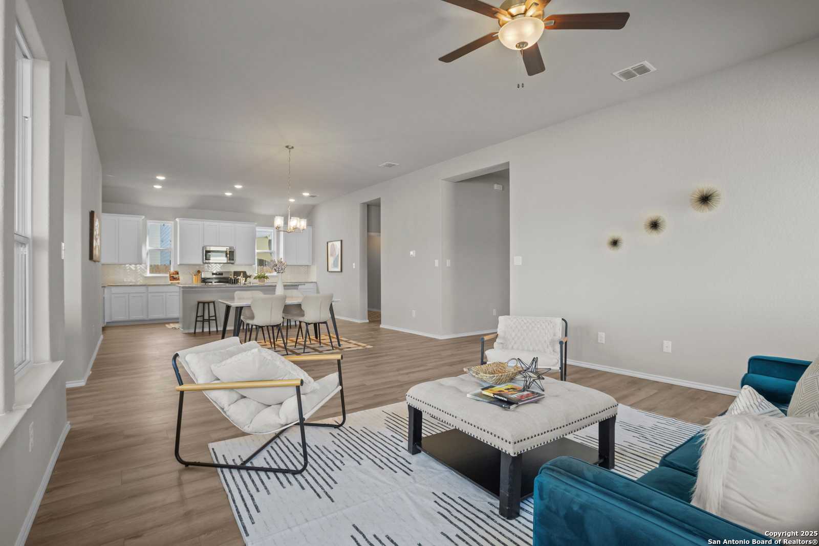 Open-concept living room with blue velvet sofa, white armchairs, hardwood floors, and adjacent kitchen in Davidson Homes The Collin B, Seguin, Texas