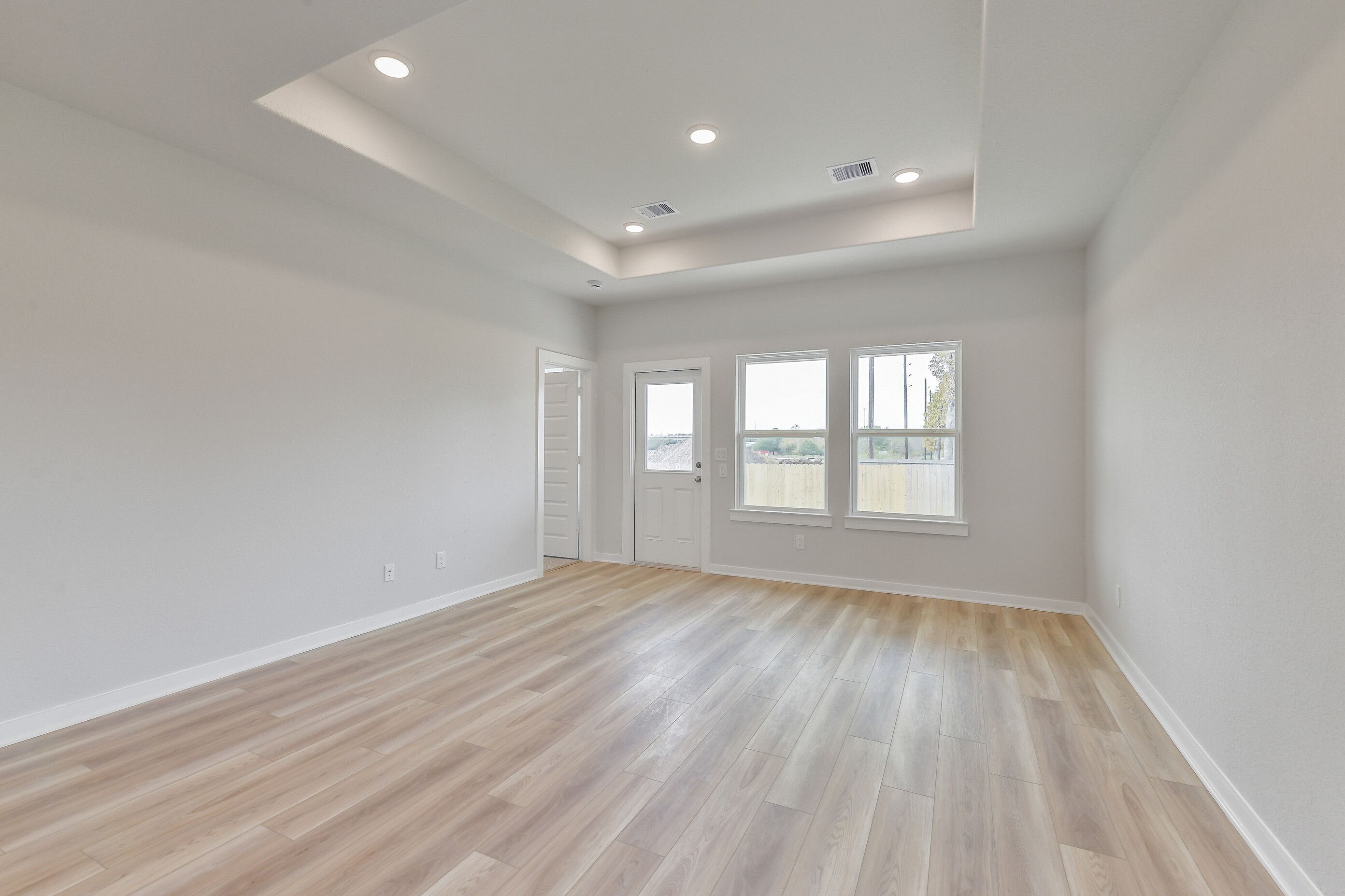Spacious living room with tray ceiling, recessed lights, large windows, and light oak laminate flooring in Davidson Homes The Colorado G, Magnolia TX