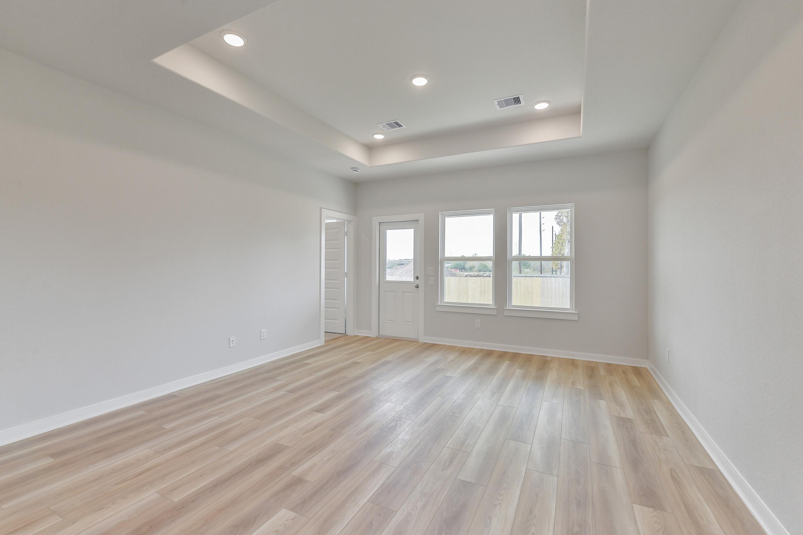 Spacious living room with tray ceiling, recessed lights, large windows, and light oak laminate flooring in Davidson Homes The Colorado G, Magnolia TX