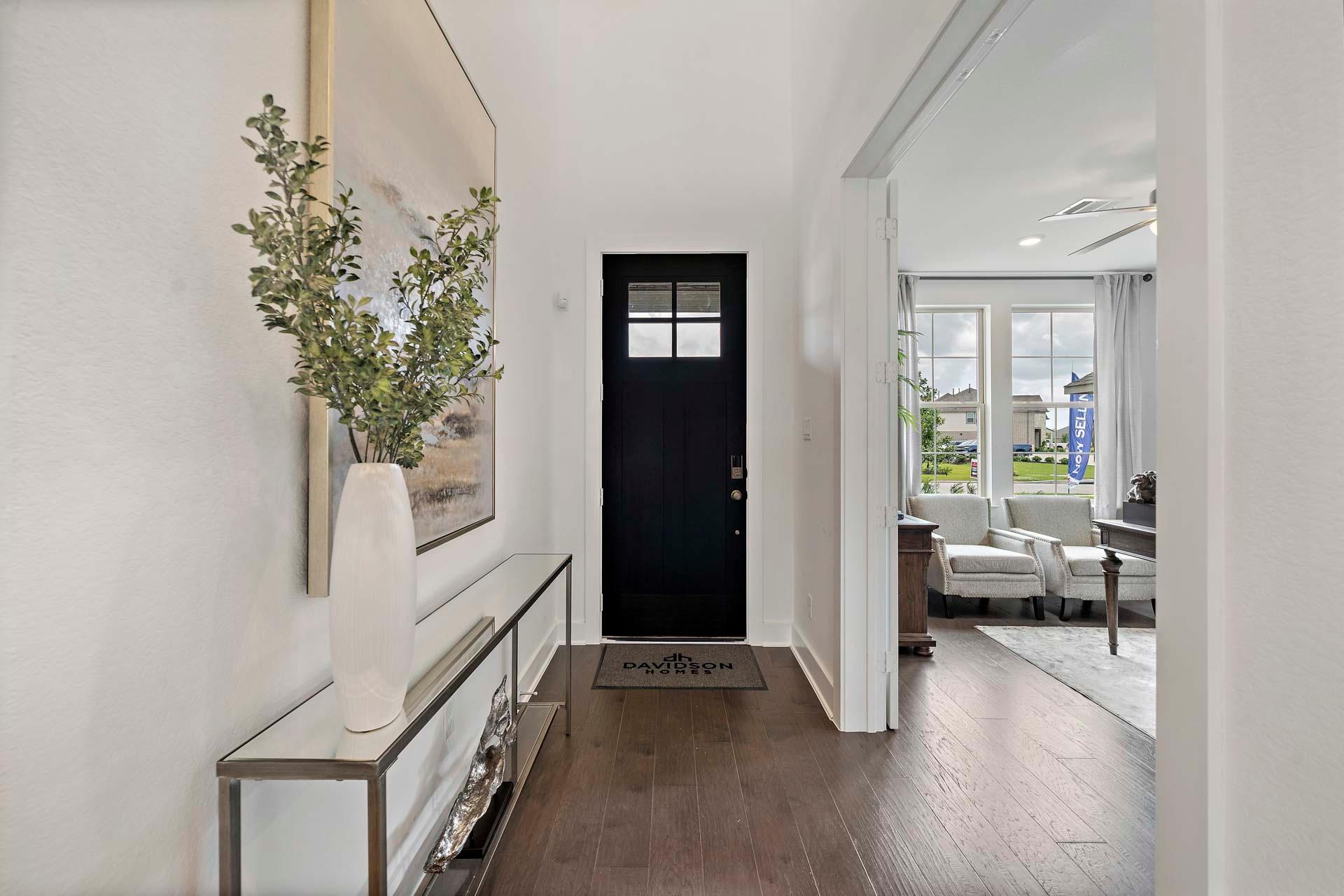 Elegant entryway at Sierra Vista in Rosharon TX with black glass door, greenery vase on console table, hardwood floors, and living room view