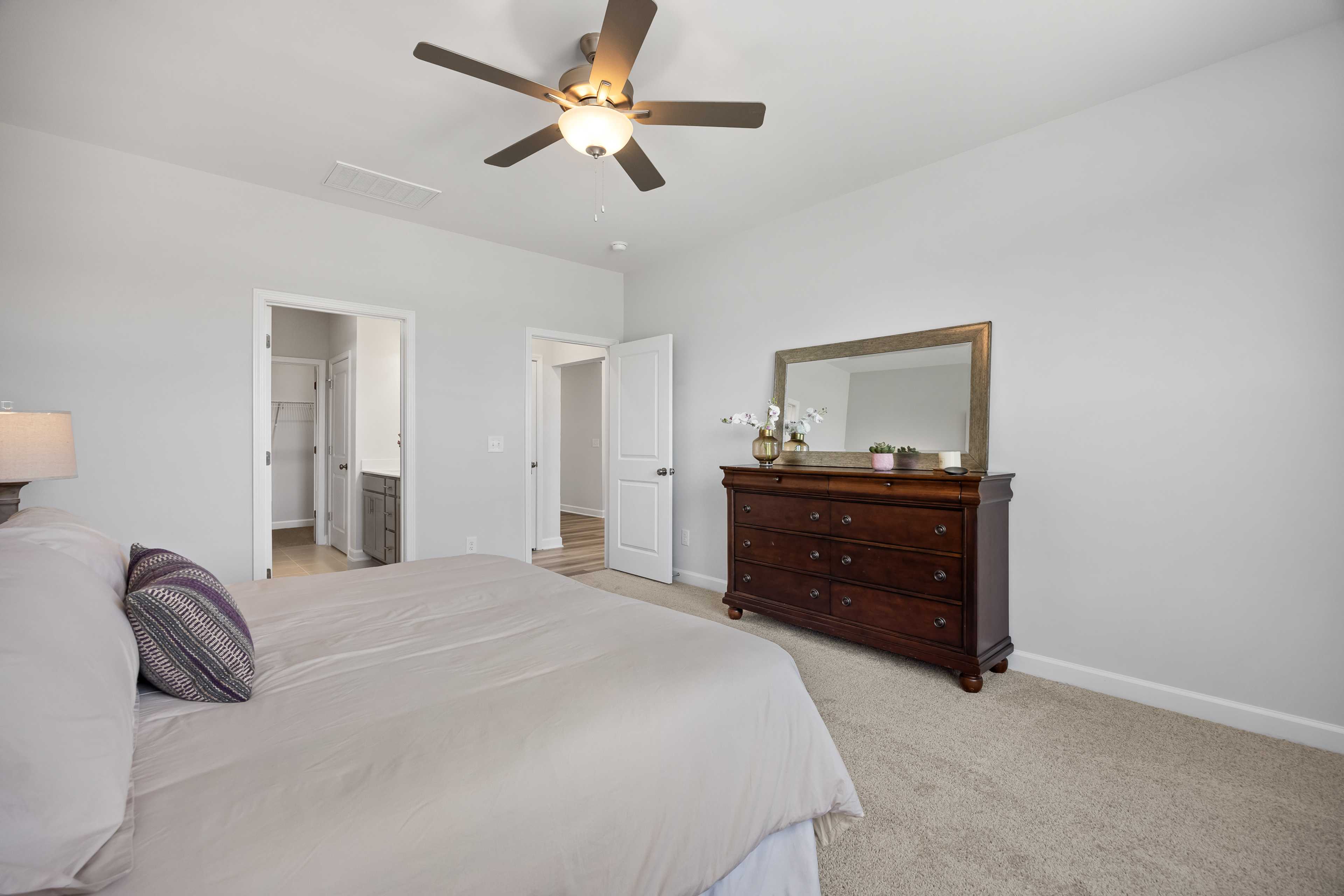 Master bedroom at Woodland Crossing in Zebulon NC with beige king bed, wooden dresser mirror, ceiling fan, and walk-in closets