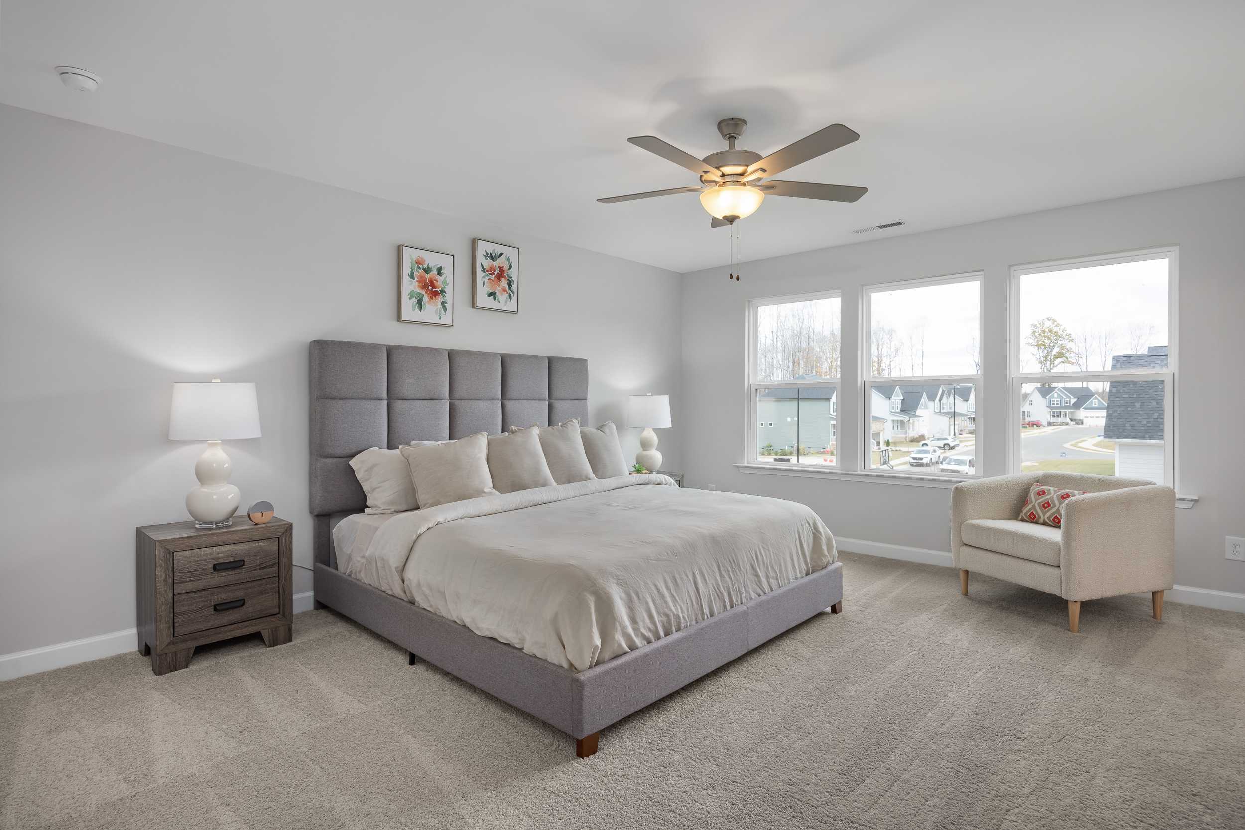 Modern primary bedroom at Enclave at Belmont in Belmont NC with gray upholstered king bed, ceiling fan, and scenic lake views
