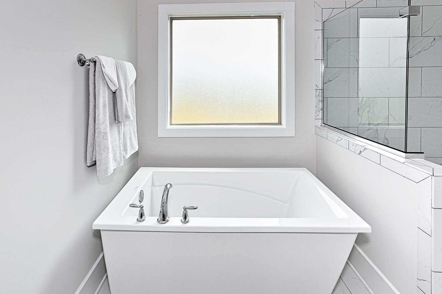Spacious master bathroom in The Finleigh with freestanding white bathtub, chrome faucets, frosted window, and subway tile shower