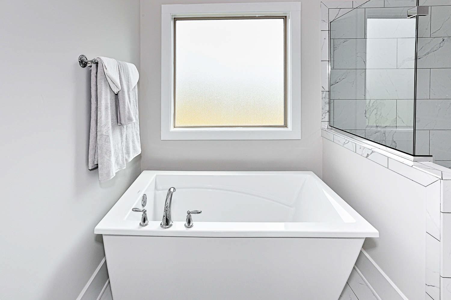 Spacious master bathroom in The Finleigh with freestanding white bathtub, chrome faucets, frosted window, and subway tile shower