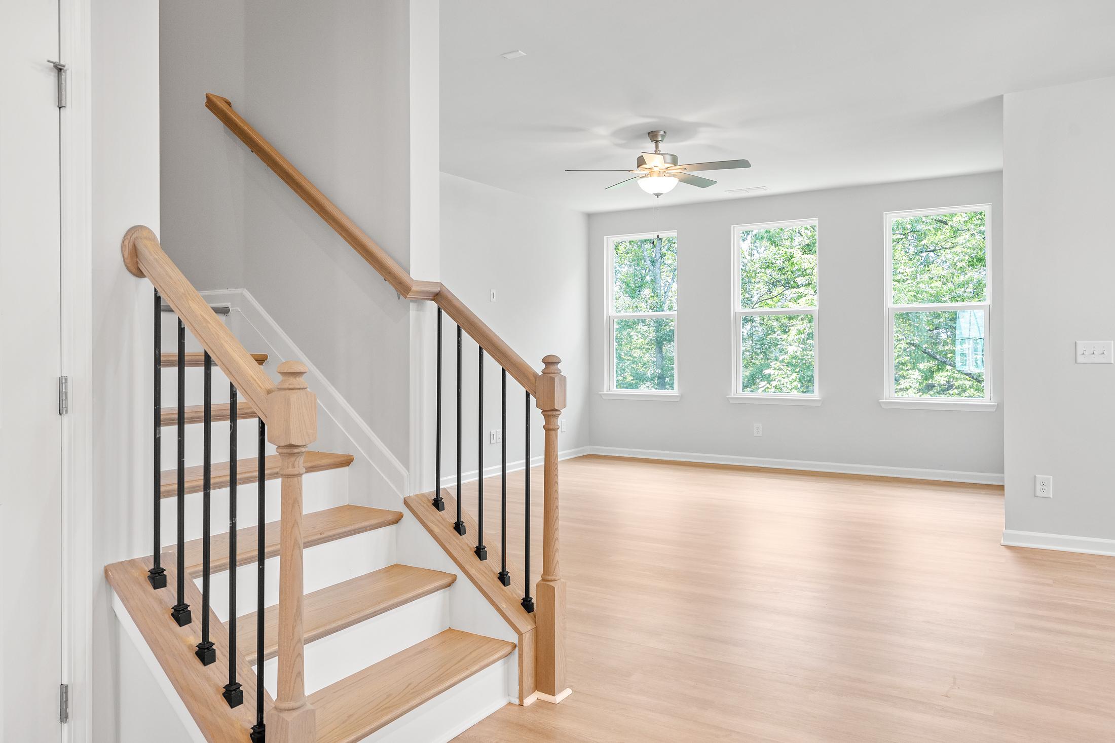 Spacious open living area in The Ash B with oak staircase, black iron railings, hardwood floors, ceiling fan, and large windows overlooking greenery