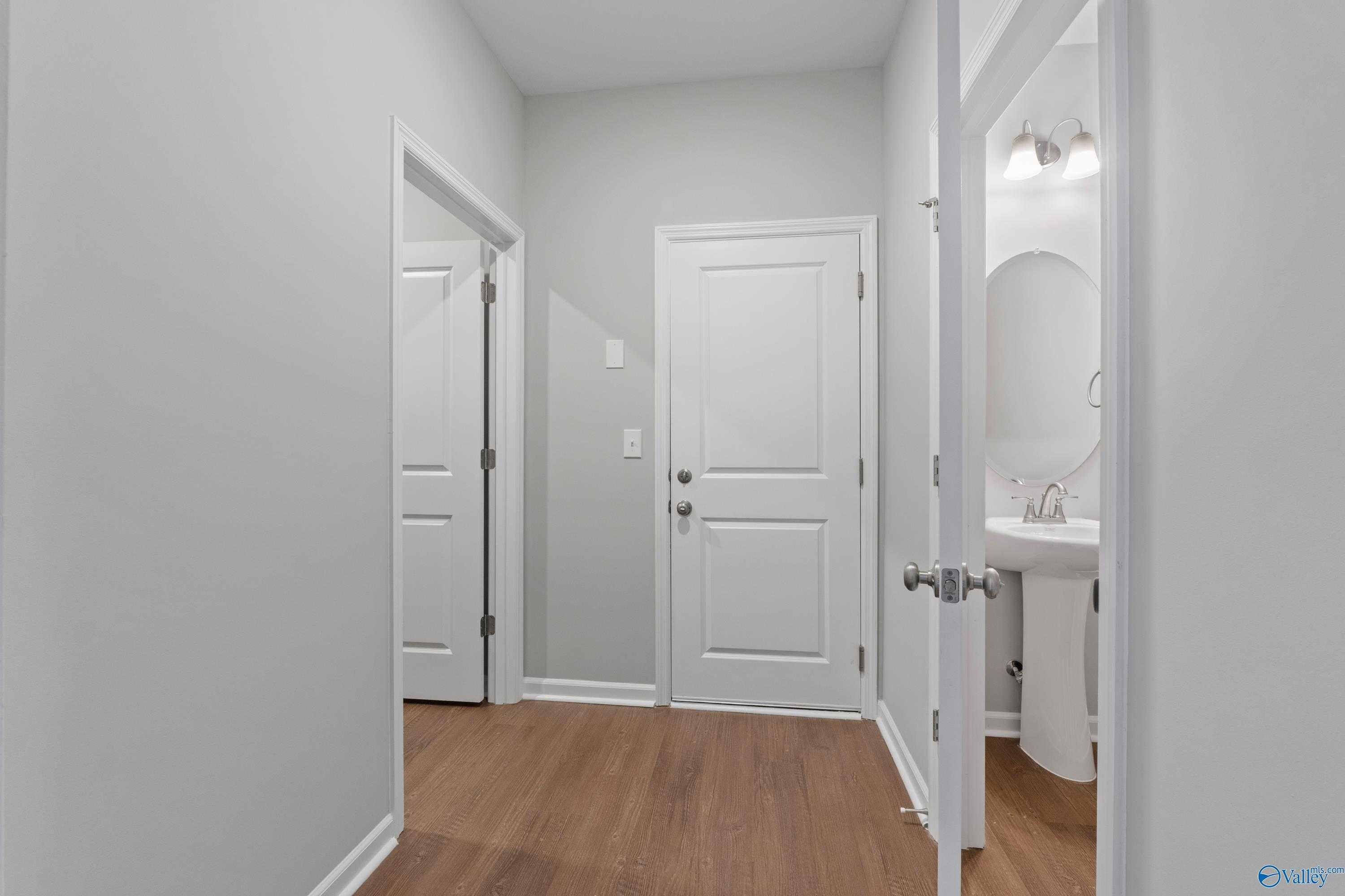 Bright hallway with light gray walls, hardwood floors, white doors, and modern bathroom featuring round mirror and pedestal sink in The Stella home