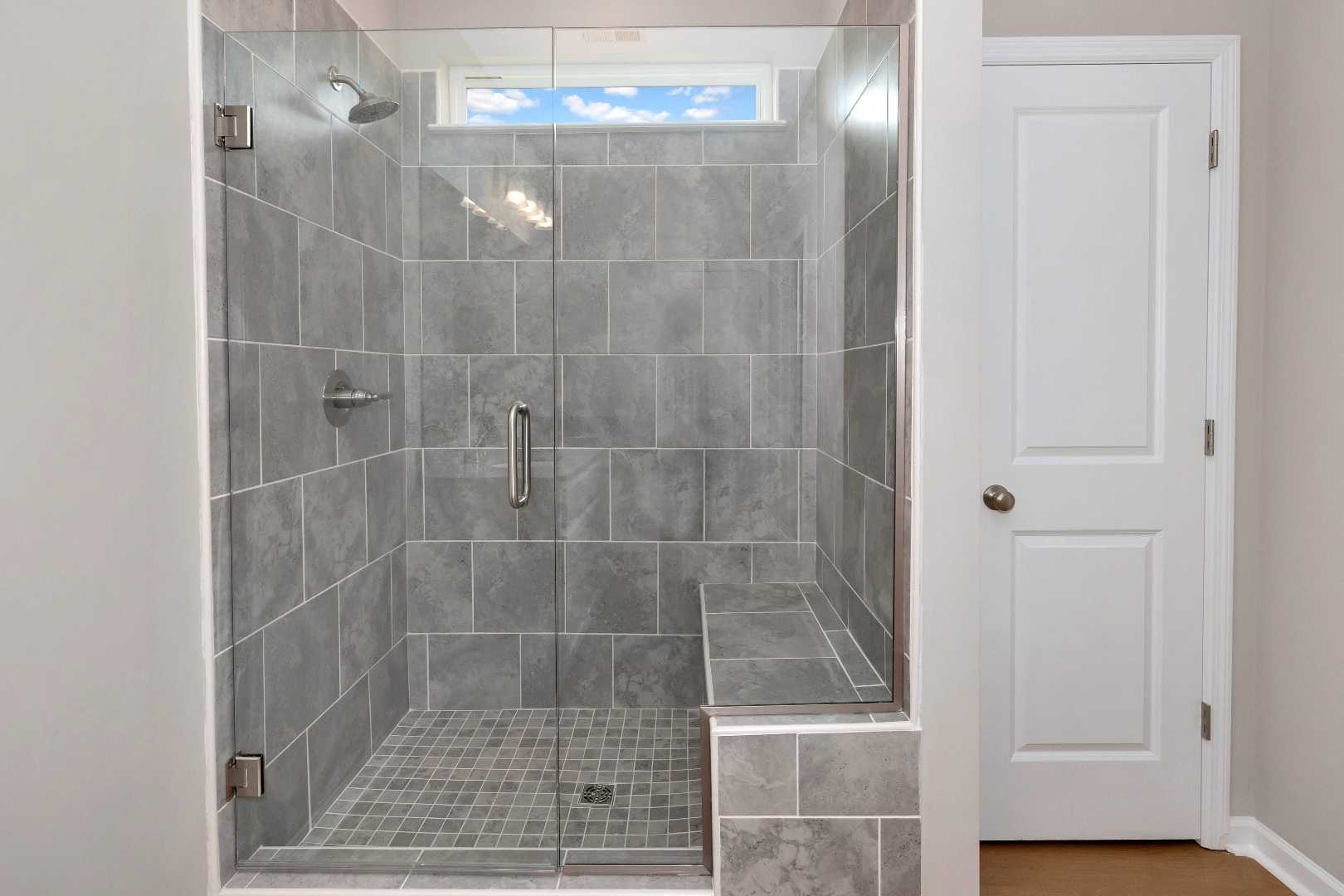 Modern walk-in shower in The Orion master bathroom with gray tile walls, glass enclosure, built-in bench, and skylight