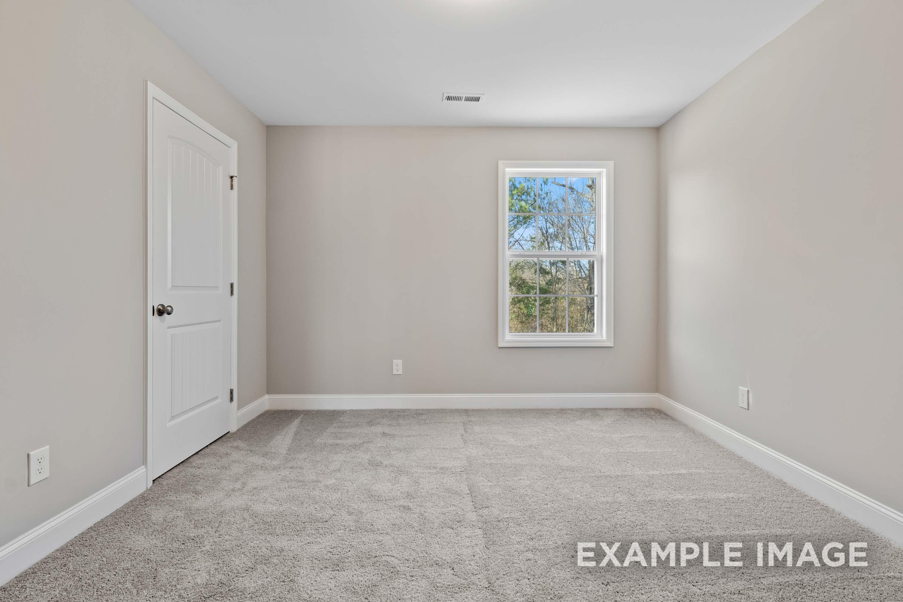 Spacious empty bedroom in The Madison B with neutral beige walls, white door, large window overlooking trees, and plush gray carpet